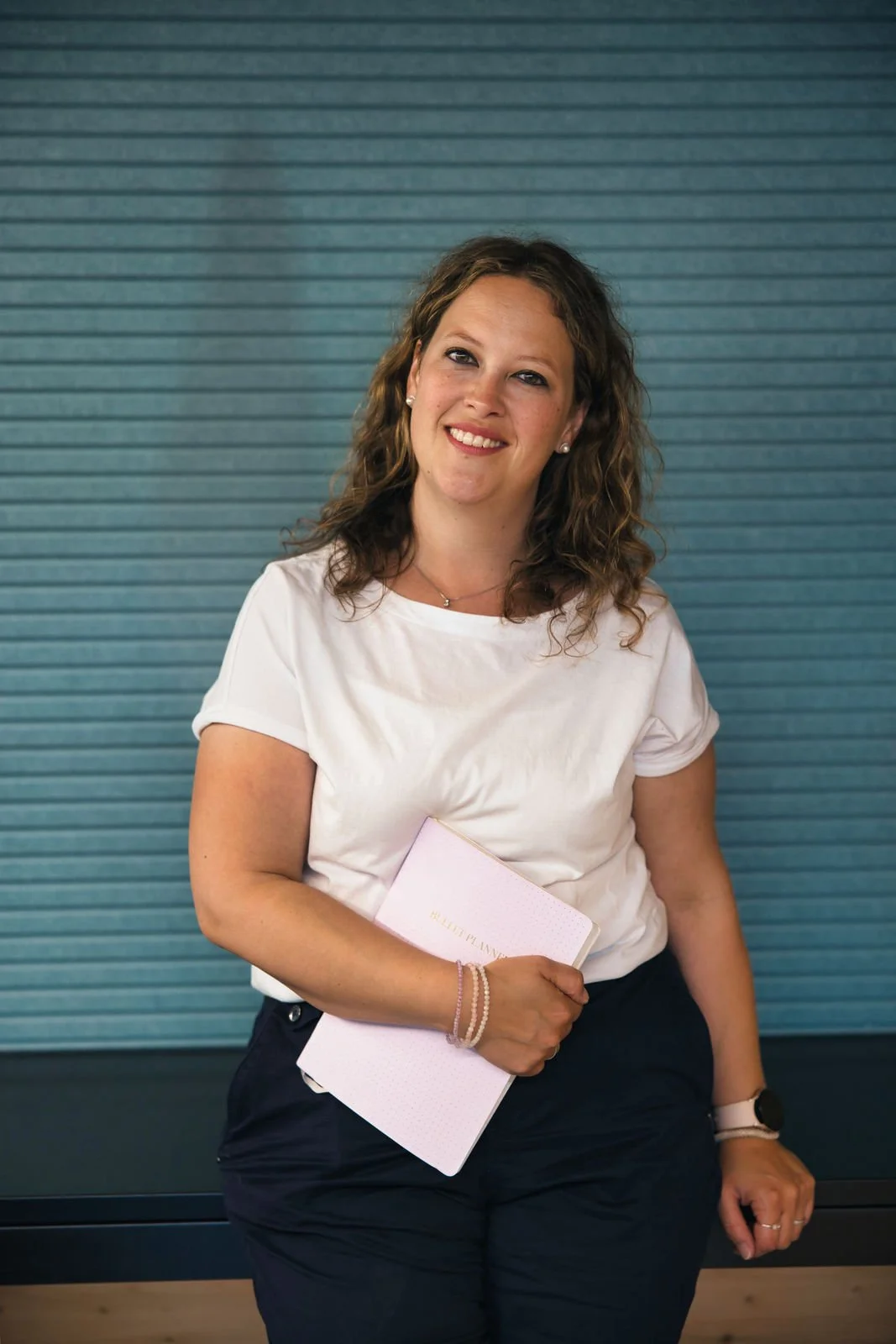 Eine lächelnde Frau mit braunen, lockigen Haaren trägt eine weiße Bluse und schwarze Hosen, hält ein Notizbuch in der Hand, steht vor einer blauen, lamellenartigen Wand.
