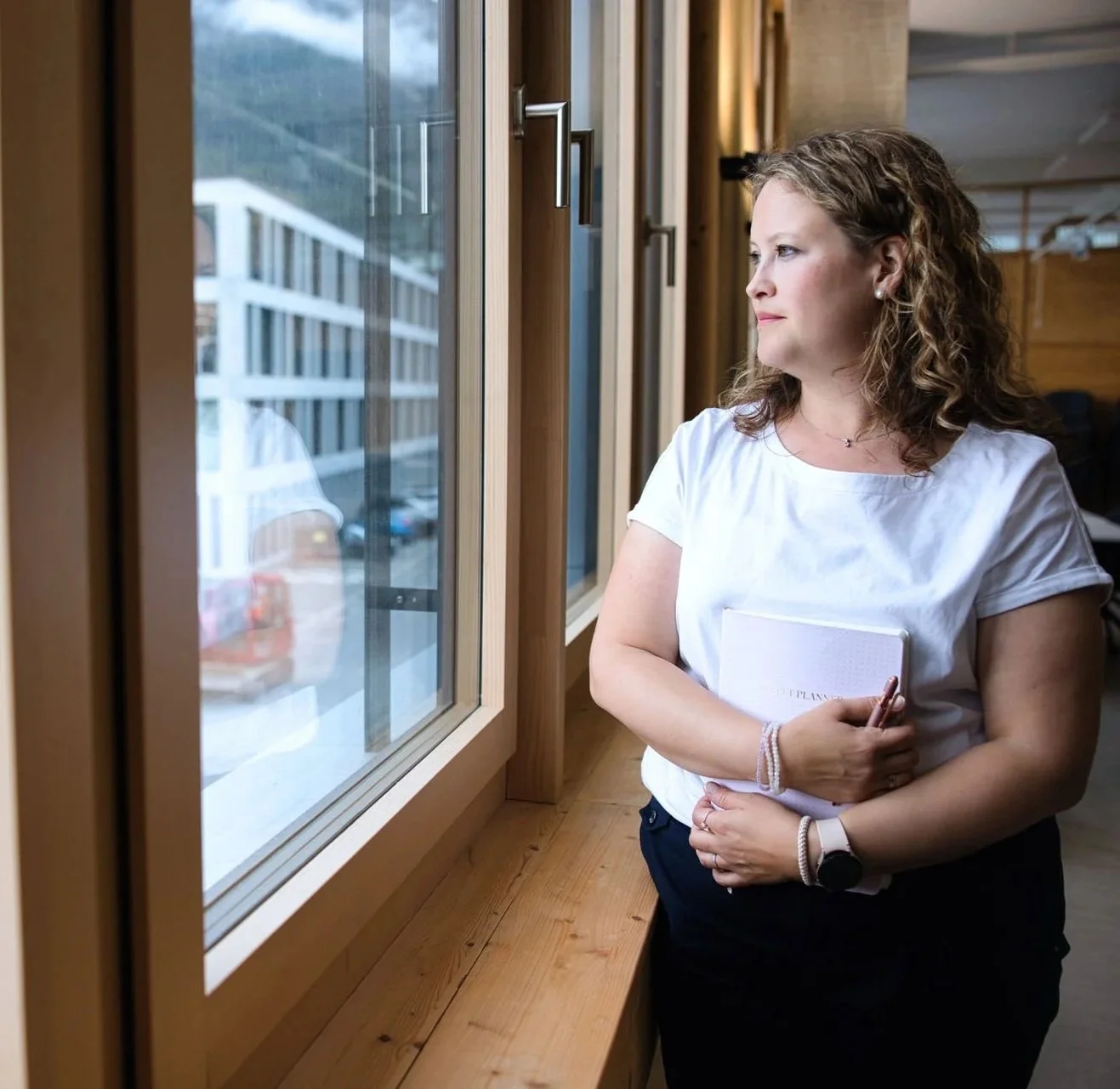 Eine Frau steht neben einem Fenster in einem Holzgebäude, hält einen Notizblock und einen Stift, und schaut nach draußen.