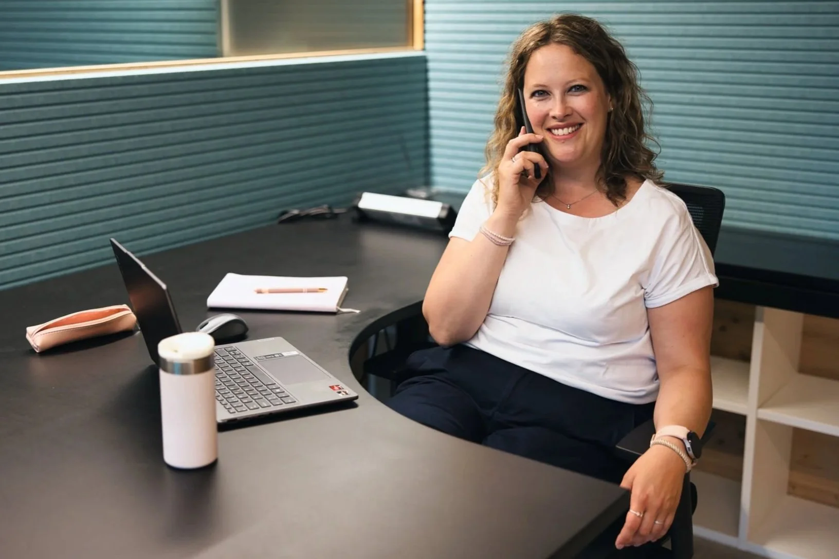 Aline Schneider, diplomierte Coachin für mentale Gesundheit, Stressprävention und Burnout-Prävention sitzt in einem Büro an einem Schreibtisch, spricht am Telefon, mit einem Laptop, Notizbuch, Stift, Wasserflasche, und dicken Pinsel auf dem Tisch.