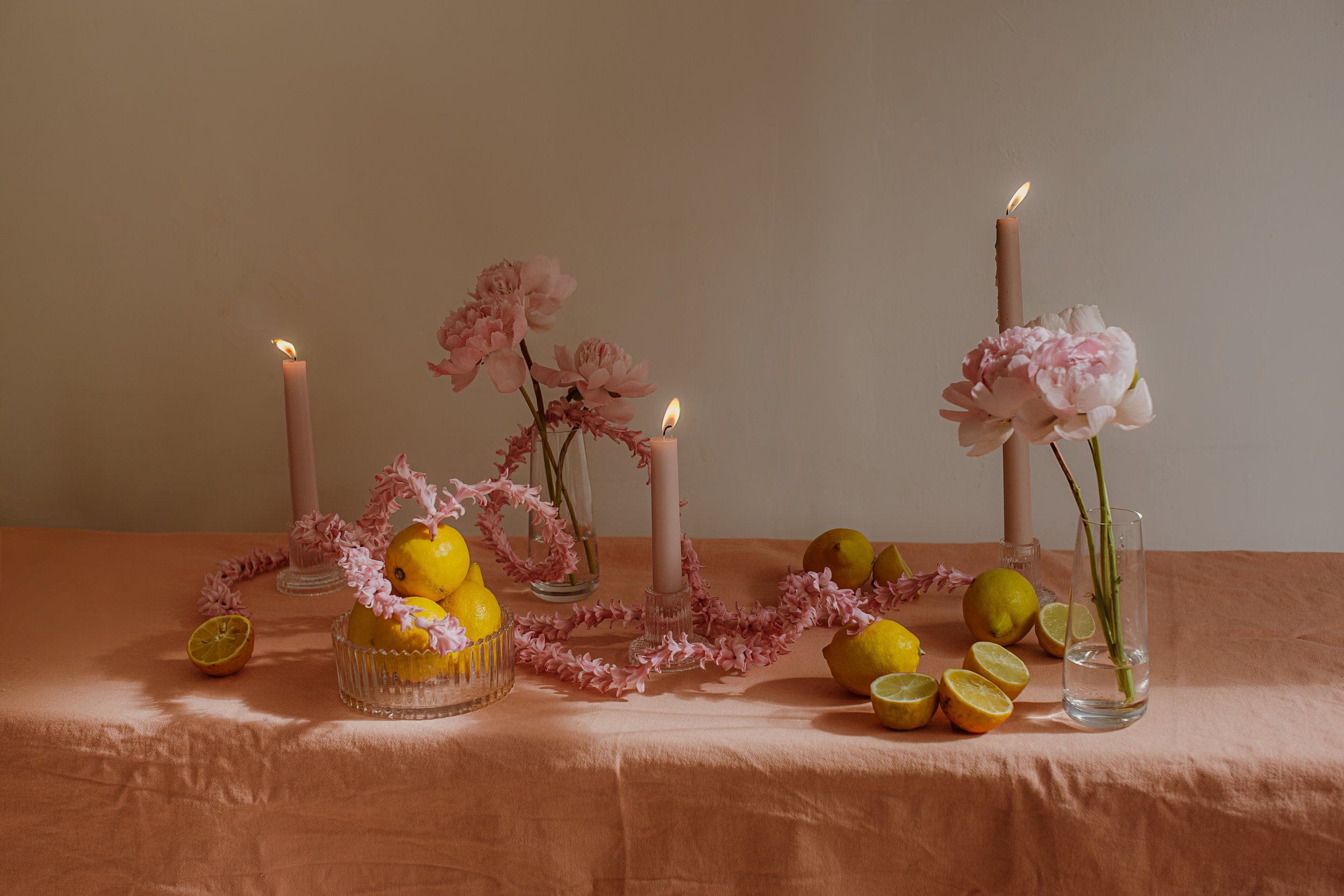 A table with pink flowers, candles, lemons, and limes on a pink tablecloth, set against a plain wall.