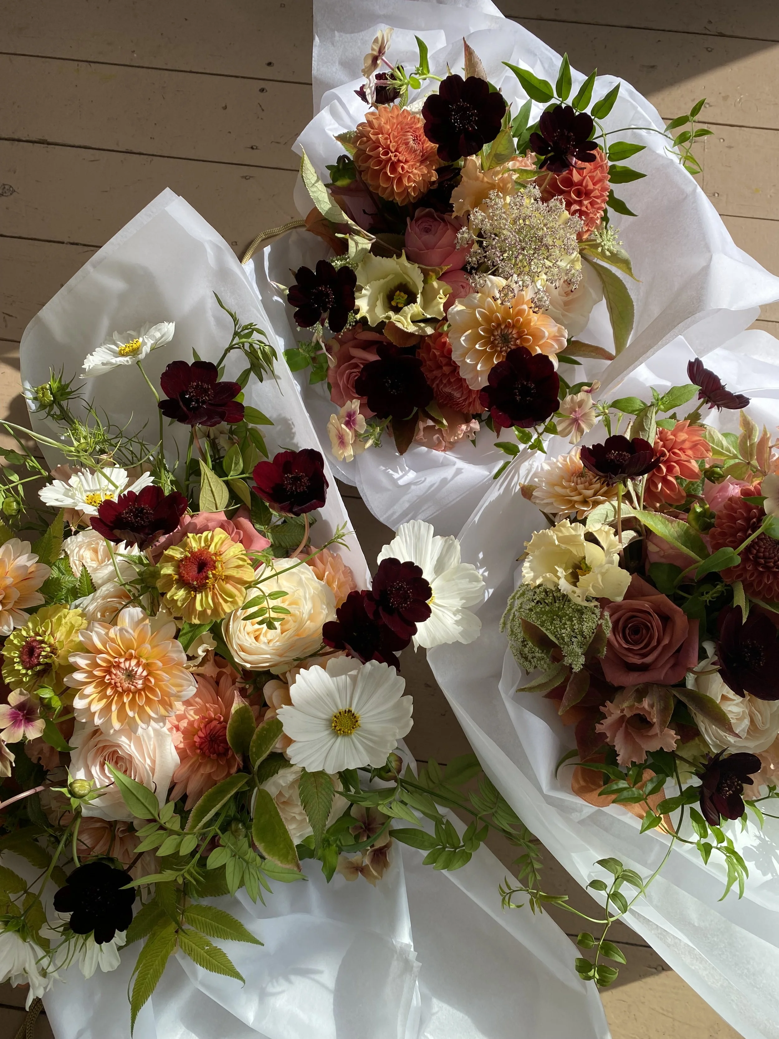 A bouquet of mixed flowers wrapped in white paper on a wooden surface under natural sunlight.