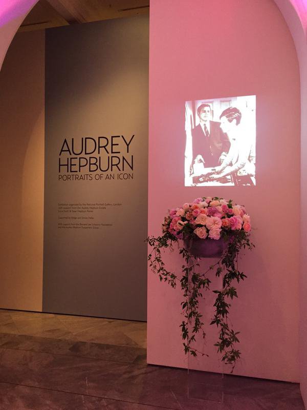 Exhibit sign titled "Audrey Hepburn: Portraits of an Icon" with a black-and-white photo of Audrey Hepburn on a pink wall, alongside a floral arrangement in a vase.