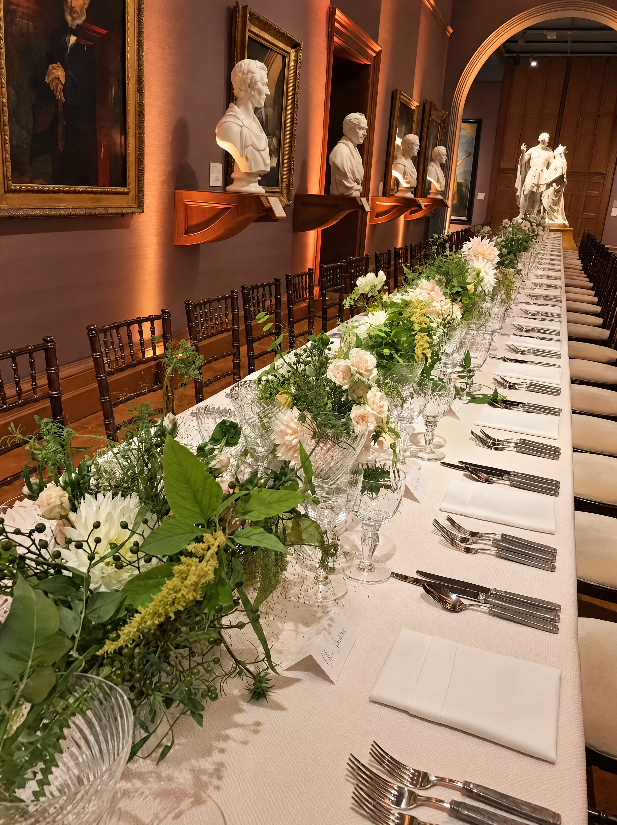 Elegant long dining table with white tablecloth, floral centerpieces, neatly arranged silverware and glassware, set for a formal event in an art-filled room with sculptures and paintings on the wall.