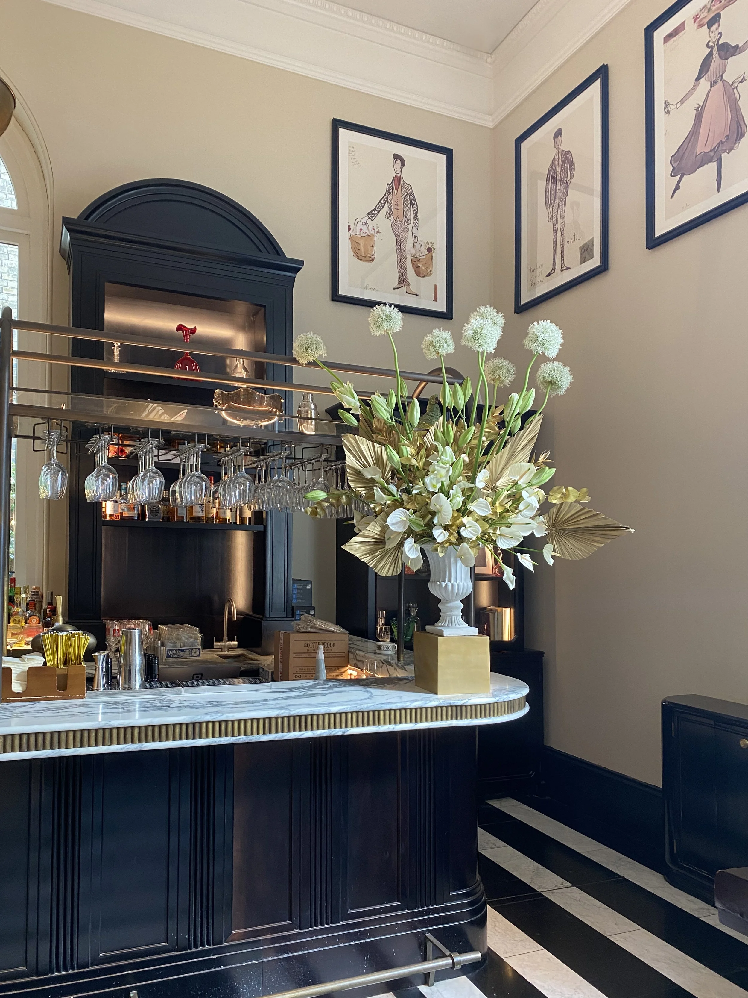 A bar area with a black wooden counter, a white marble top, and hanging wine glasses. A large floral arrangement with white flowers and green foliage is on the counter. Three framed fashion sketches hang on the beige wall behind the bar.