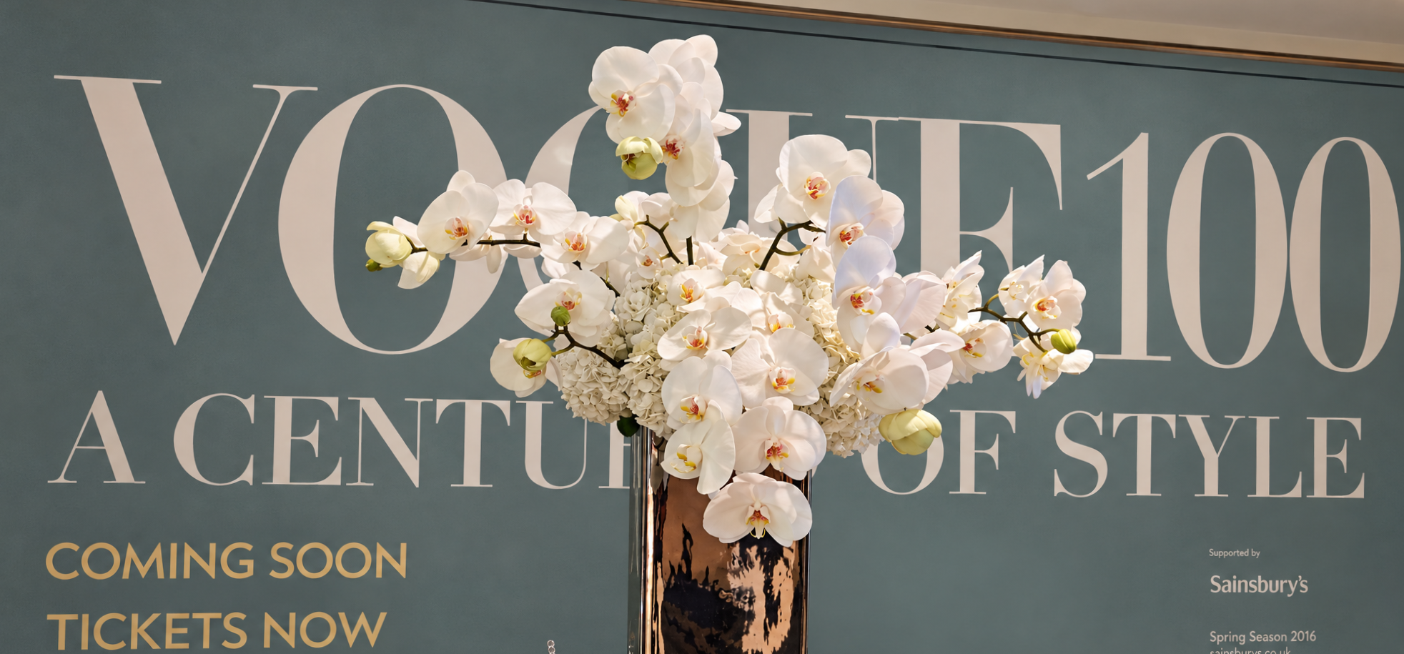 A large floral arrangement of white orchids and hydrangeas in a tall, dark vase in front of a teal background with the text 'VOCAL' and other words.