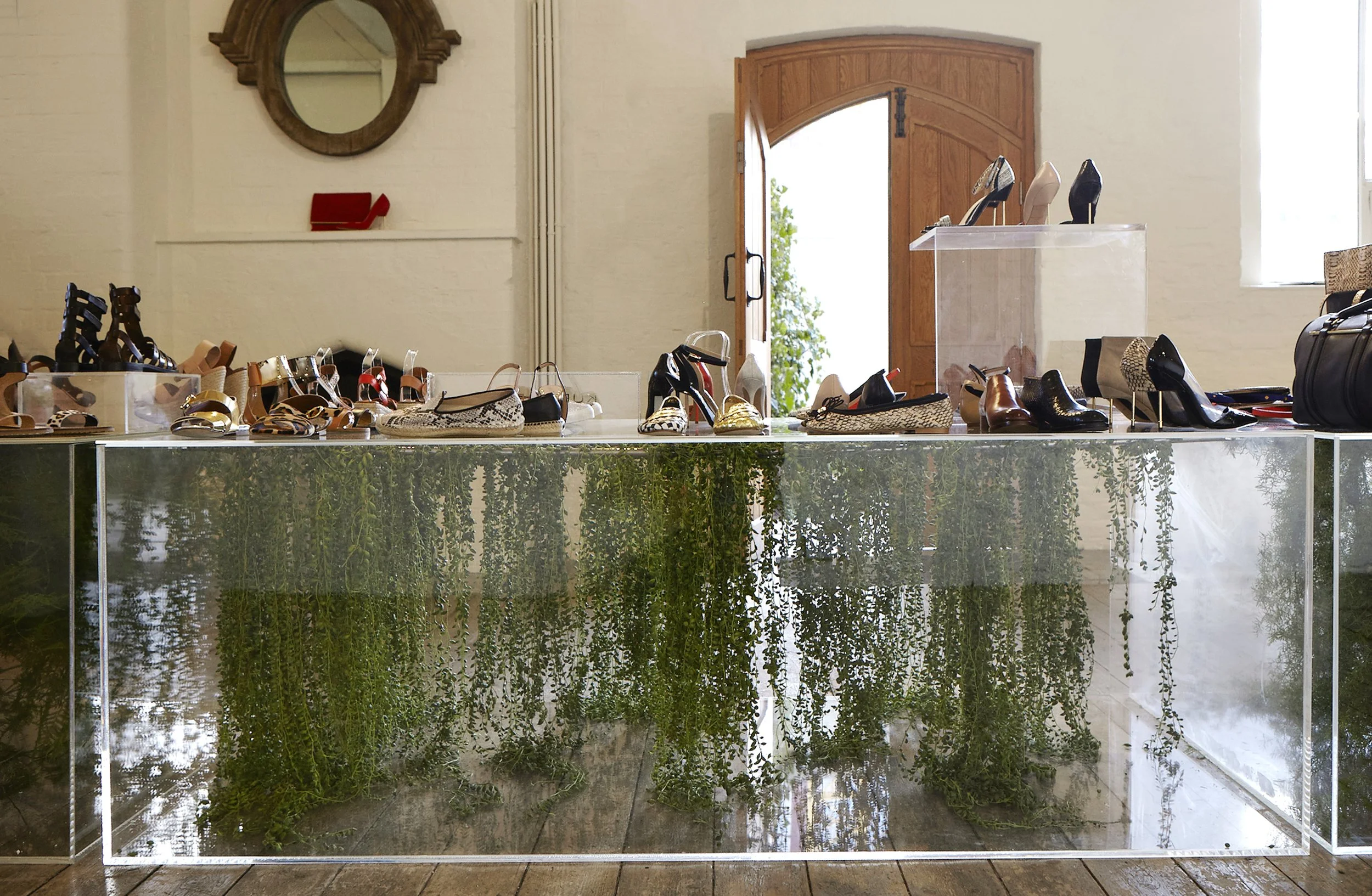 Display of various pairs of high-heeled shoes arranged on a table with a glass enclosure filled with green plants underneath, in a room with a wooden door, a mirror, and cream-colored walls.