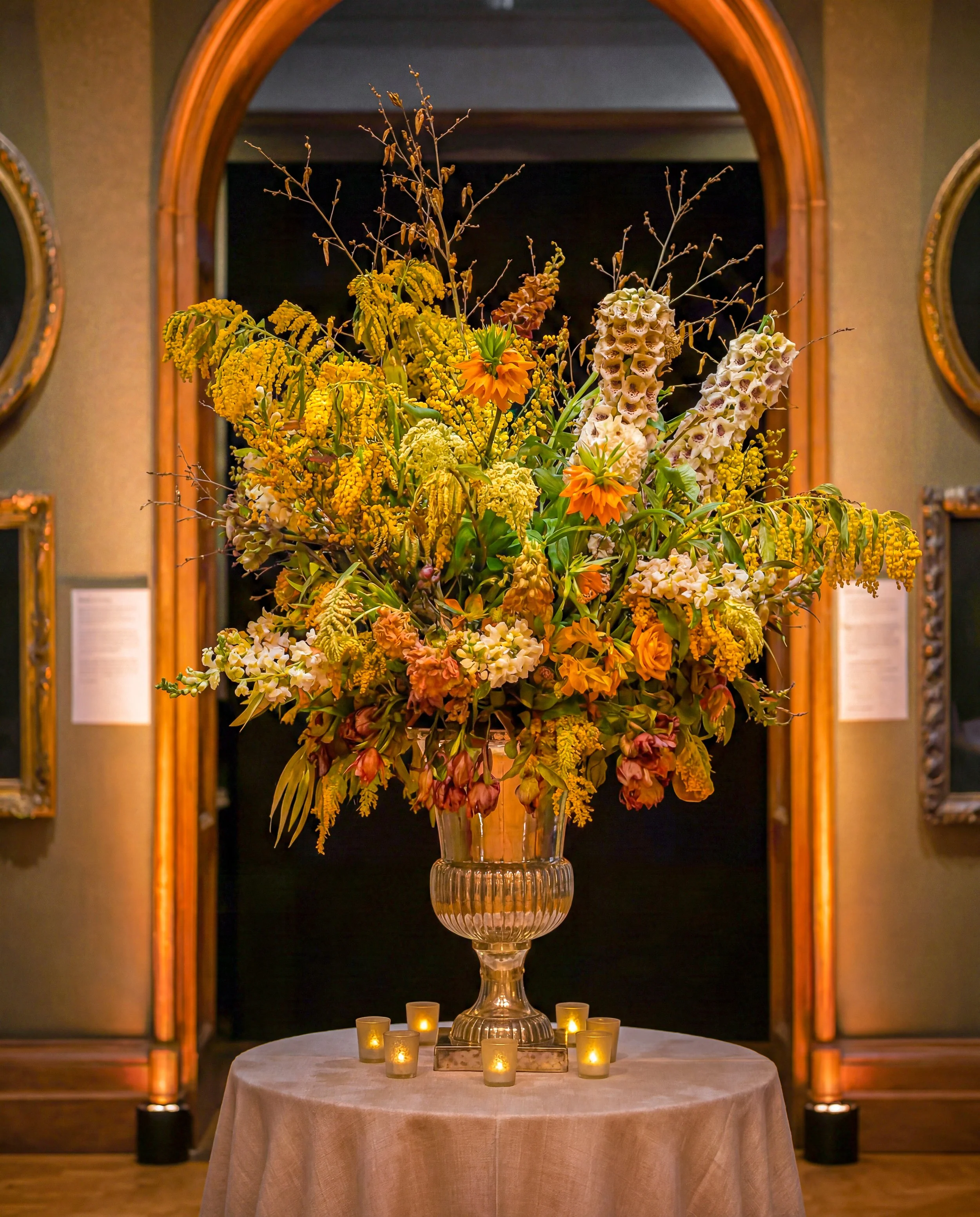 A large floral arrangement in a glass vase on a table, surrounded by small lit candles, with a wooden archway and framed mirrors in the background.