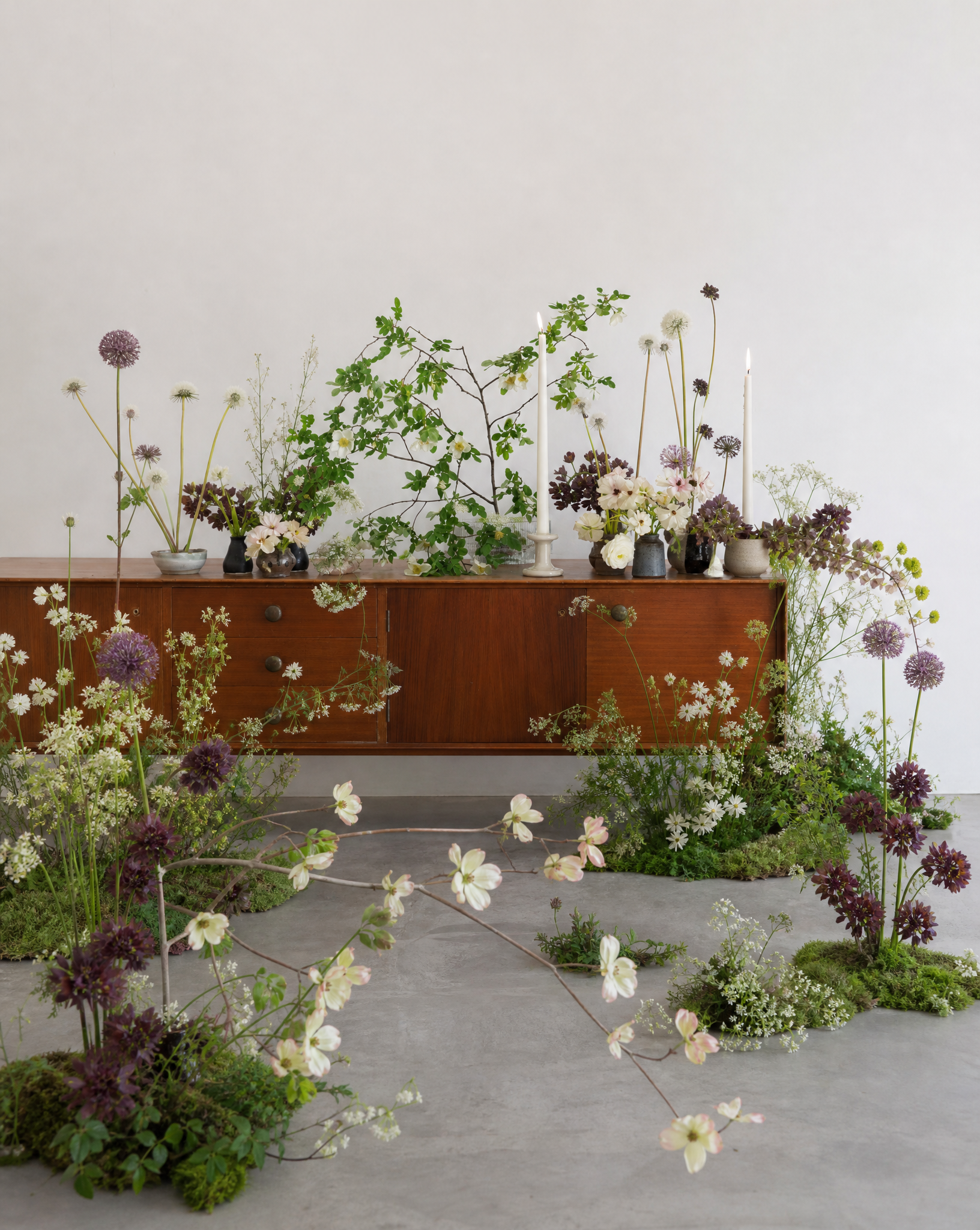 Arrangement of various flowers and greenery in vases on a wooden sideboard with additional flowers and plants on the floor in front.