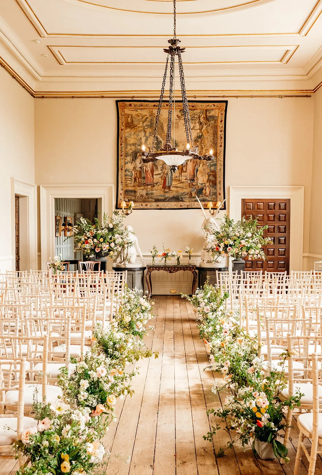 An elegant indoor wedding venue decorated with floral arrangements, with an aisle lined with white chairs and a floral altar at the front, featuring large sculptures of animals and a tapestry on the wall.
