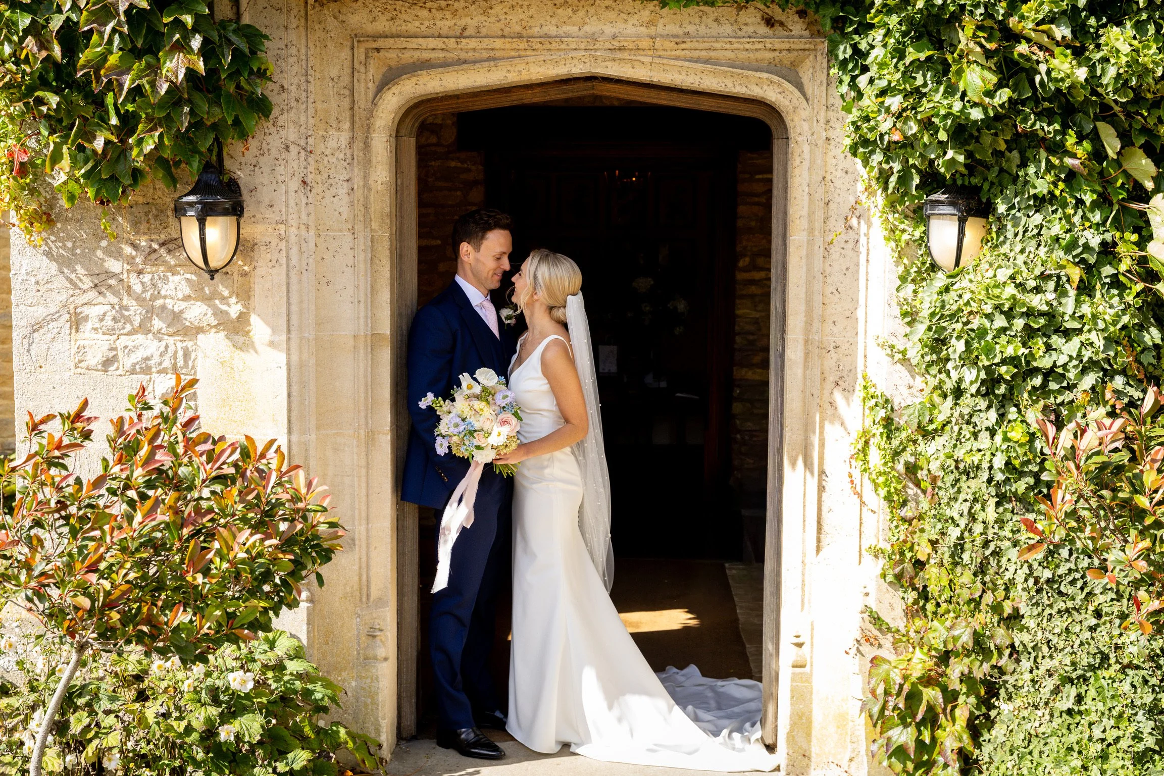 A bride and groom embrace under a stone archway, framed by green foliage, during their wedding.