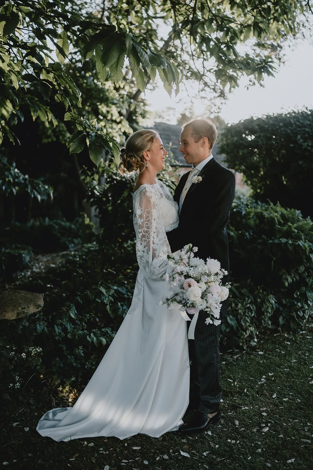 A bride and groom stand close together outdoors, smiling and looking into each other's eyes, surrounded by greenery and trees with sunlight filtering through the leaves. The bride wears a white lace wedding dress and holds a bouquet of flowers, while