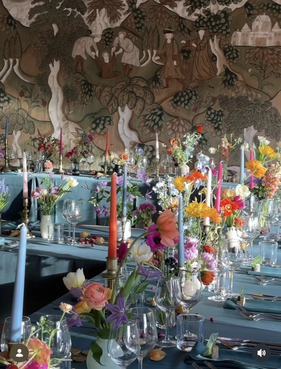 A decorated banquet table set with colorful flowers, candles, glassware, and cutlery, with a large ornate mural of trees and people in vintage attire on the wall behind.