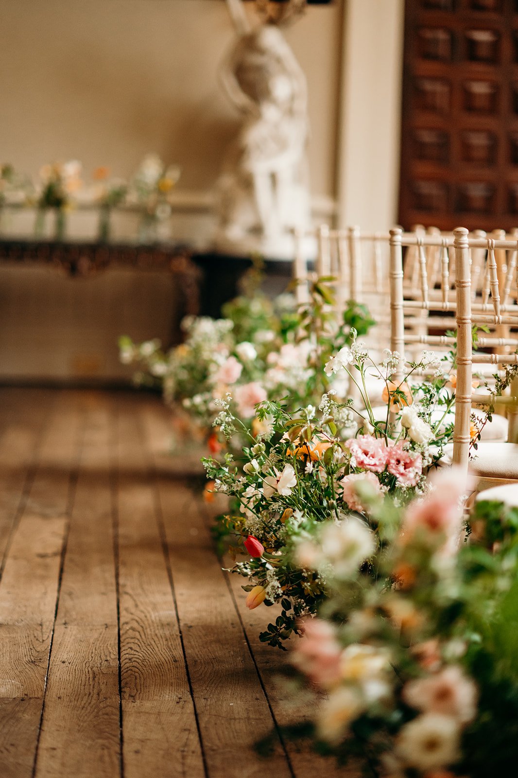 Rows of beige chairs decorated with pink, white, and orange flowers at a formal event with wooden flooring and an elegant background.