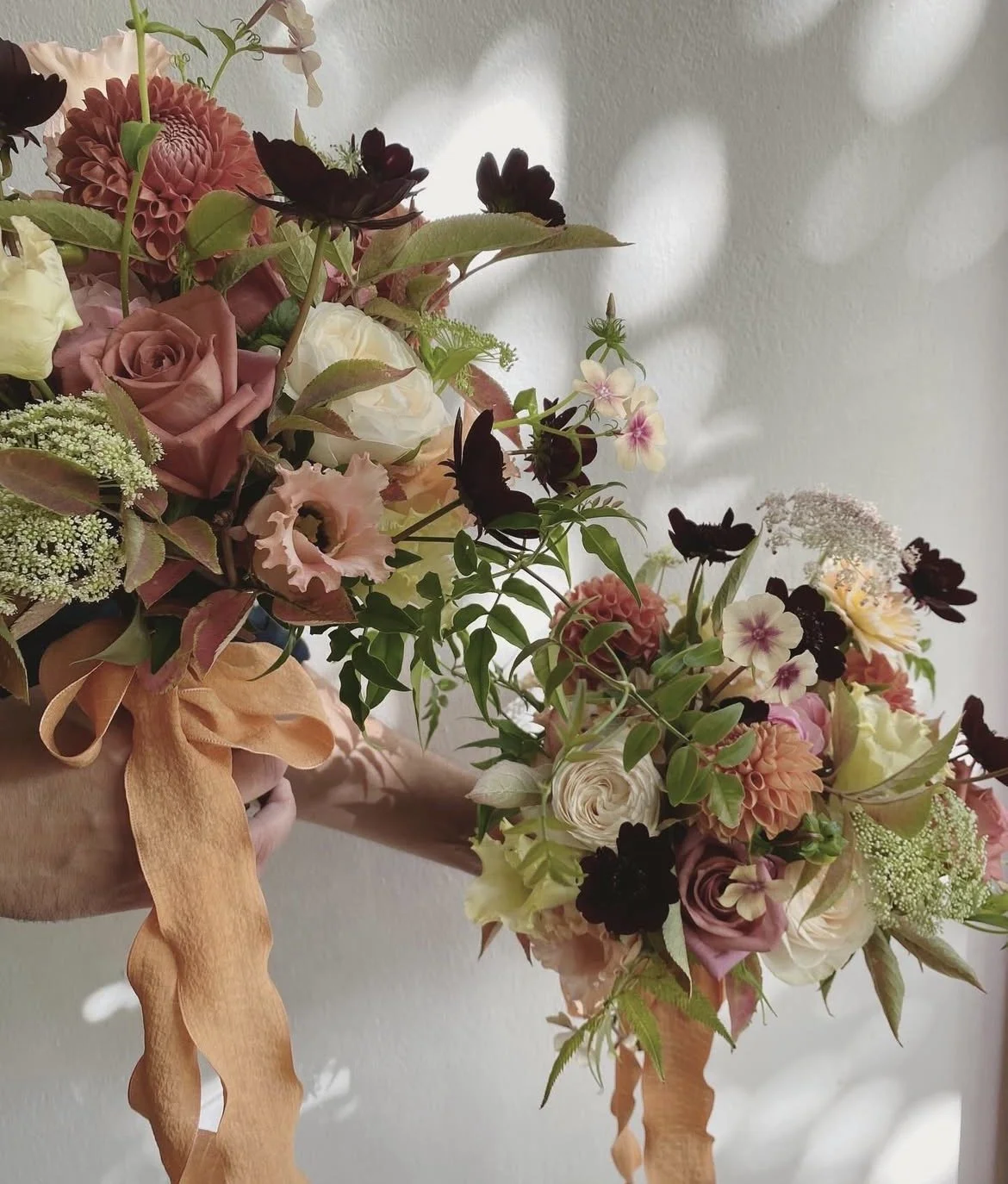 A bouquet of pink, cream, white, and dark purple flowers with green leaves, wrapped with a peach-colored ribbon, against a light-colored background.