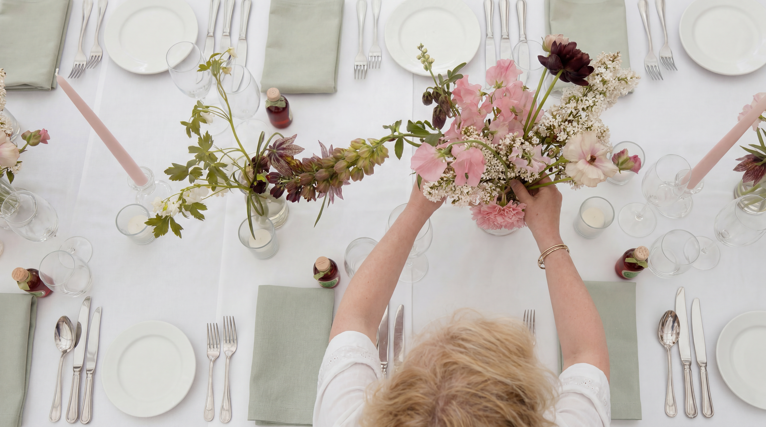 Overhead view of a person arranging pink and white flowers in a vase on a dining table set for a formal dinner with white plates, silver cutlery, light green napkins, and candles.