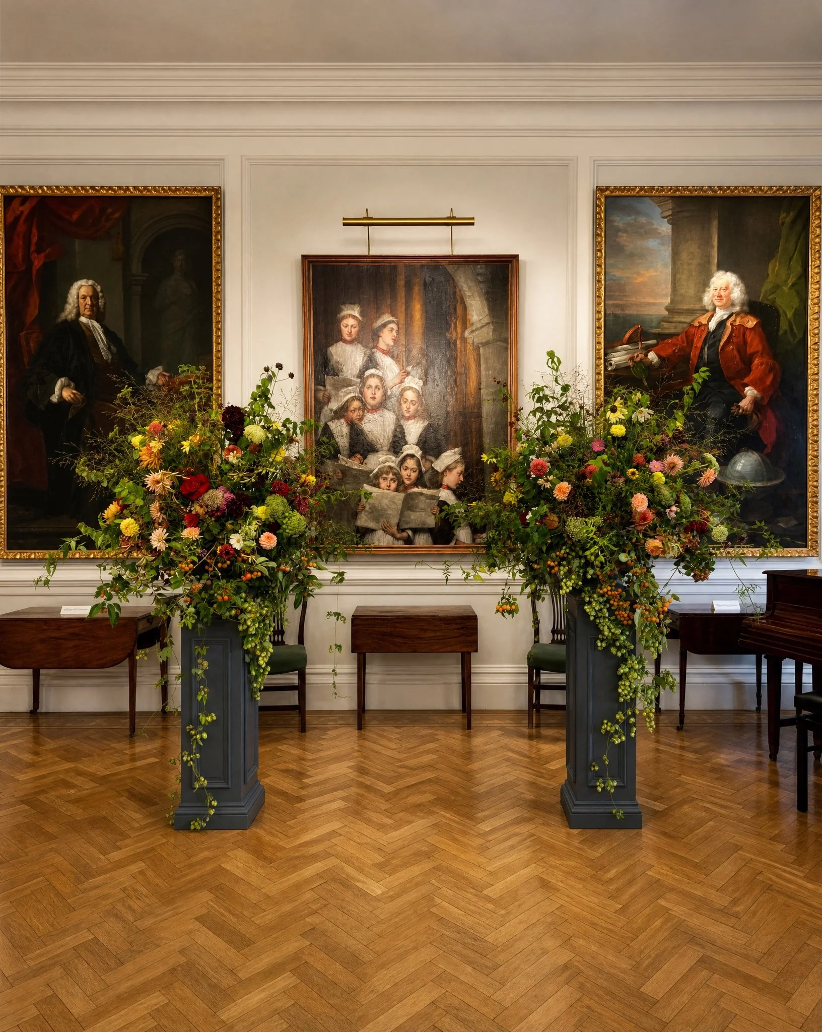 A museum or gallery interior with three large portrait paintings on the wall, two of elderly men and one of a group of children and women, with floral arrangements on two pedestals in front of the paintings, wooden flooring, and white decorative wall