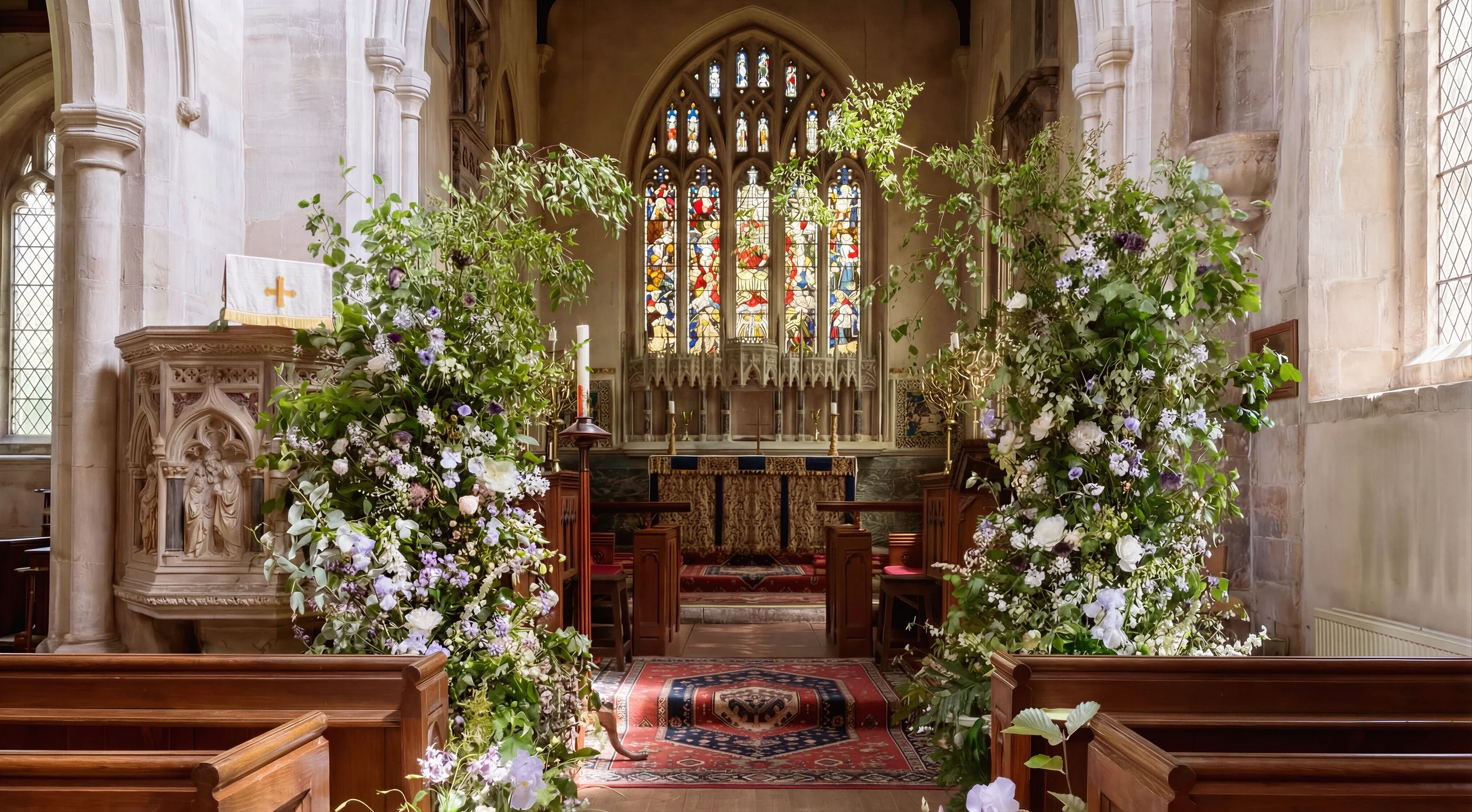 Interior of a church altar decorated with greenery and flowers, featuring stained glass windows and wooden pews.