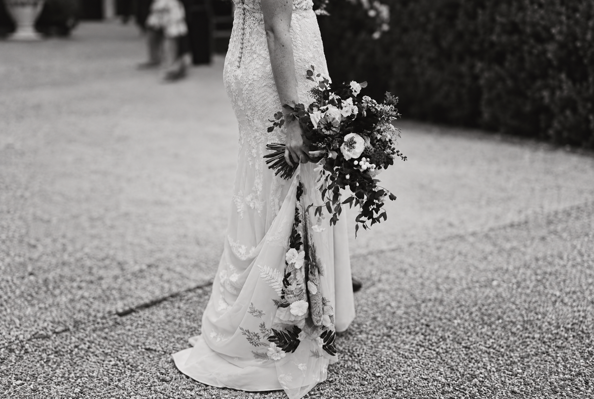 A bride in a lace wedding dress holding a bouquet of flowers, standing outdoors on a gravel path with people in the background.