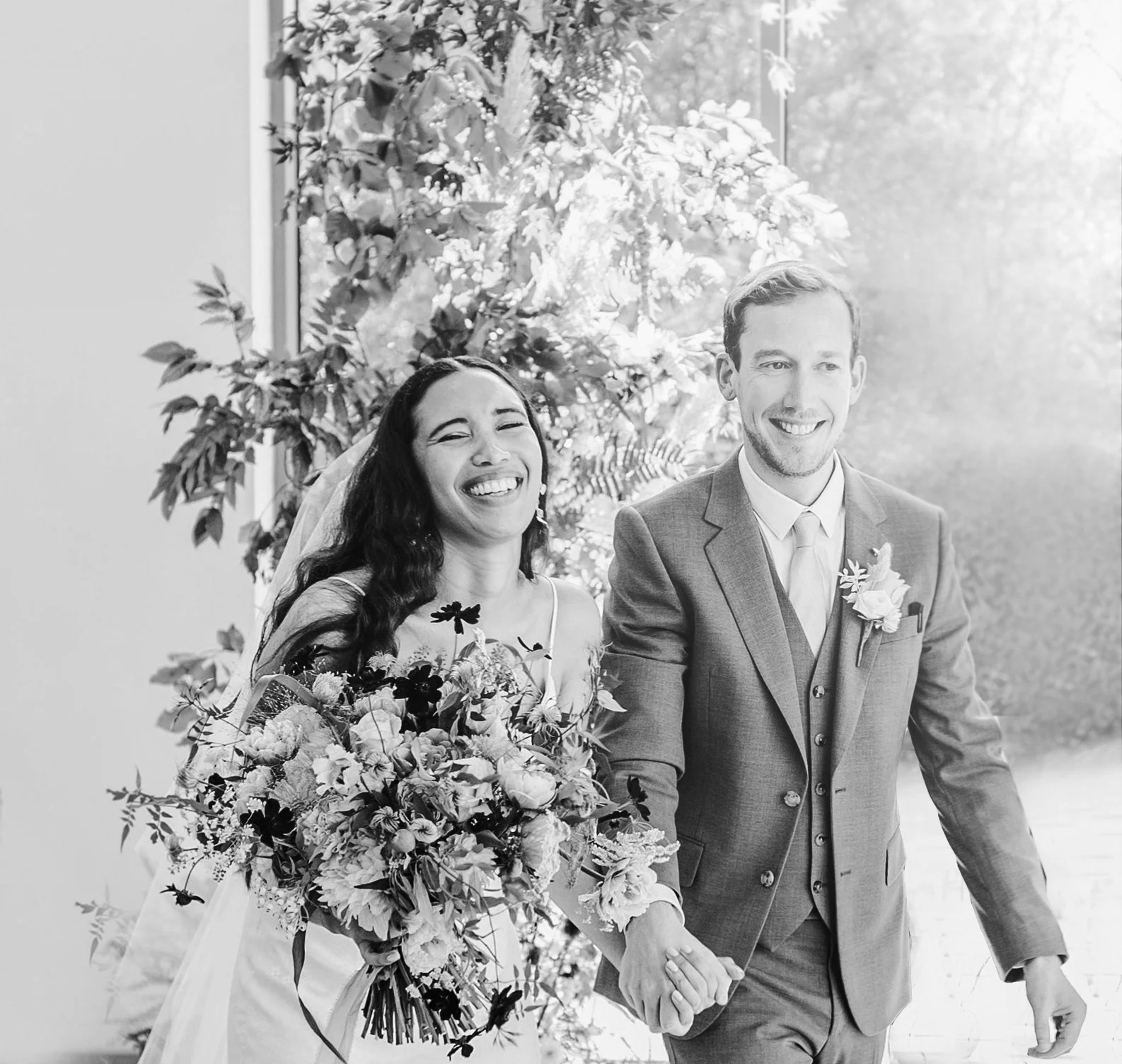 A smiling bride and groom holding hands during their wedding ceremony, surrounded by flowers and greenery, outdoors in natural light.