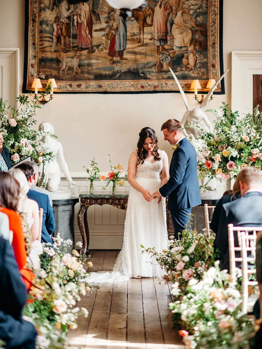 A couple getting married during a wedding ceremony in a decorated indoor venue with floral arrangements and a large tapestry on the wall.