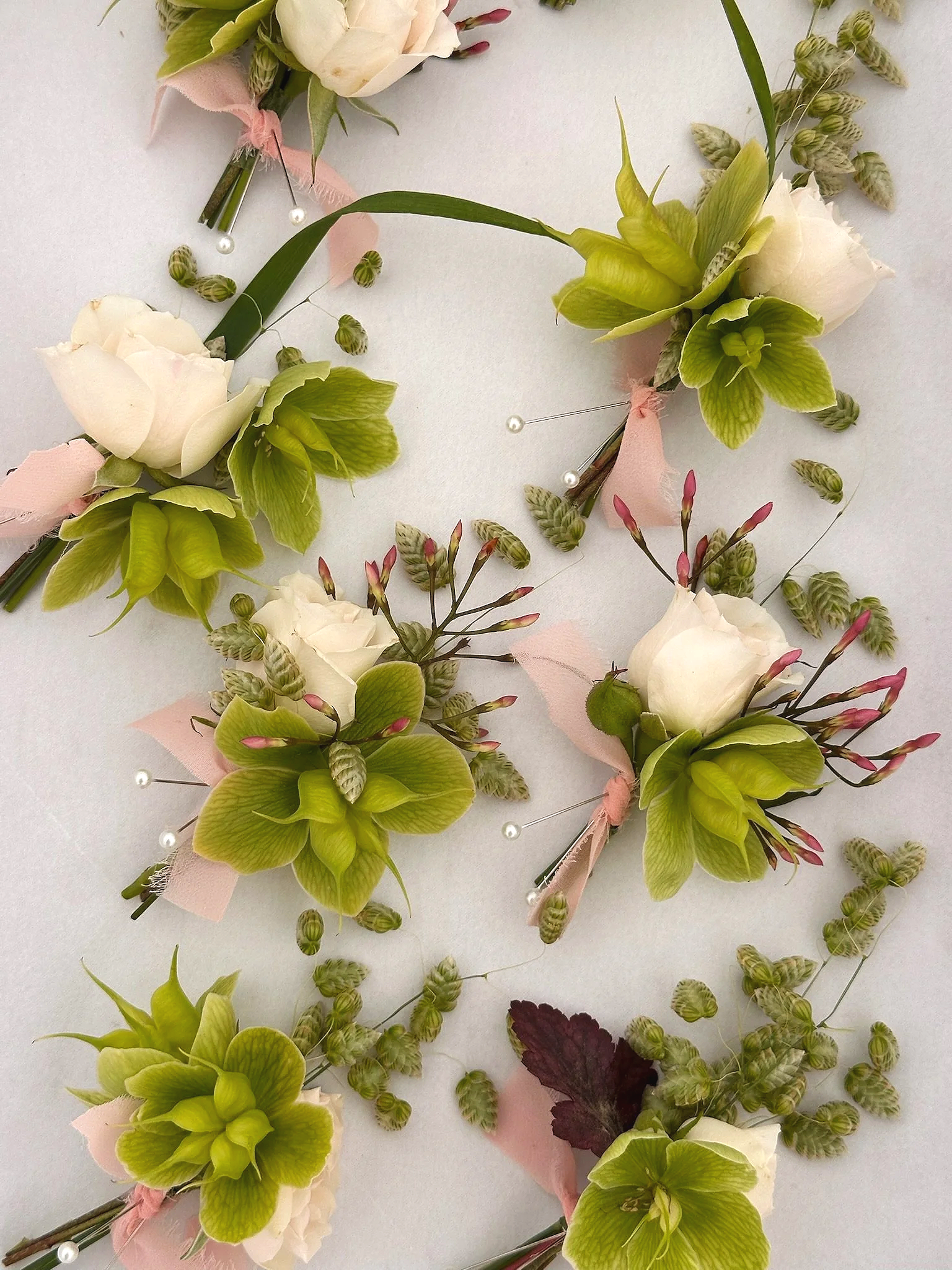Arrangement of floral corsages or boutonnieres with white and green flowers, pink ribbons, and pearl pins on a white surface.