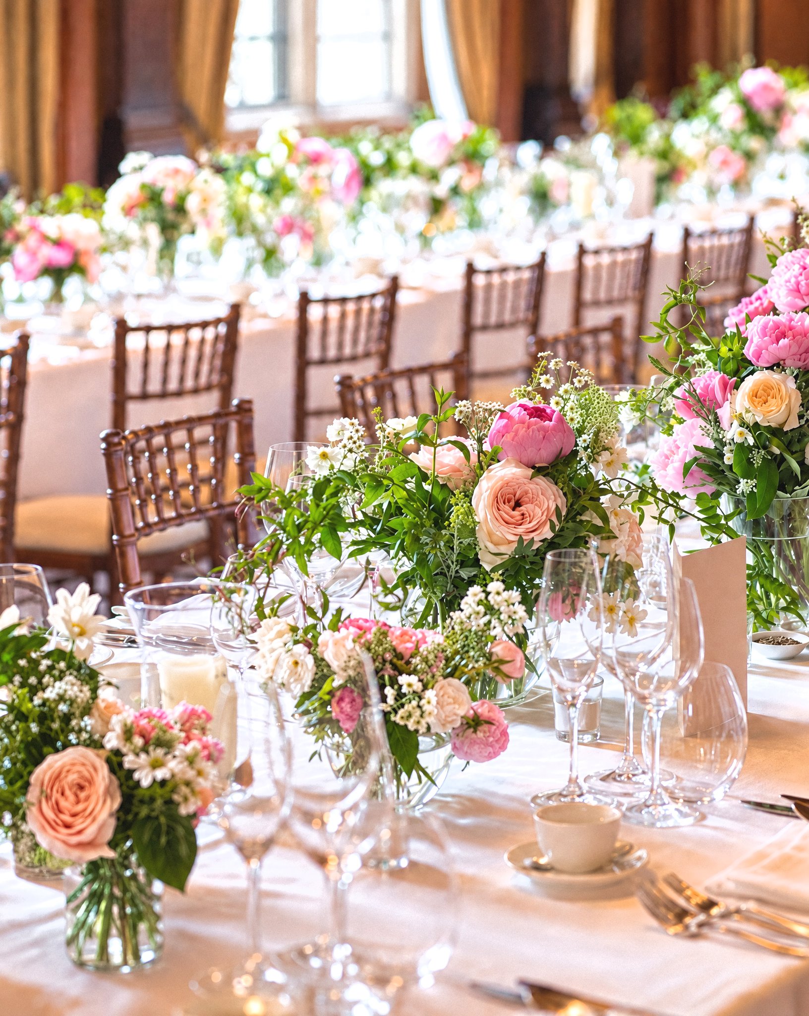 A decorated banquet table set for a wedding or special event, with floral centerpieces featuring pink, peach, and white flowers and glassware, in a rustic wooden venue with windows and curtains.