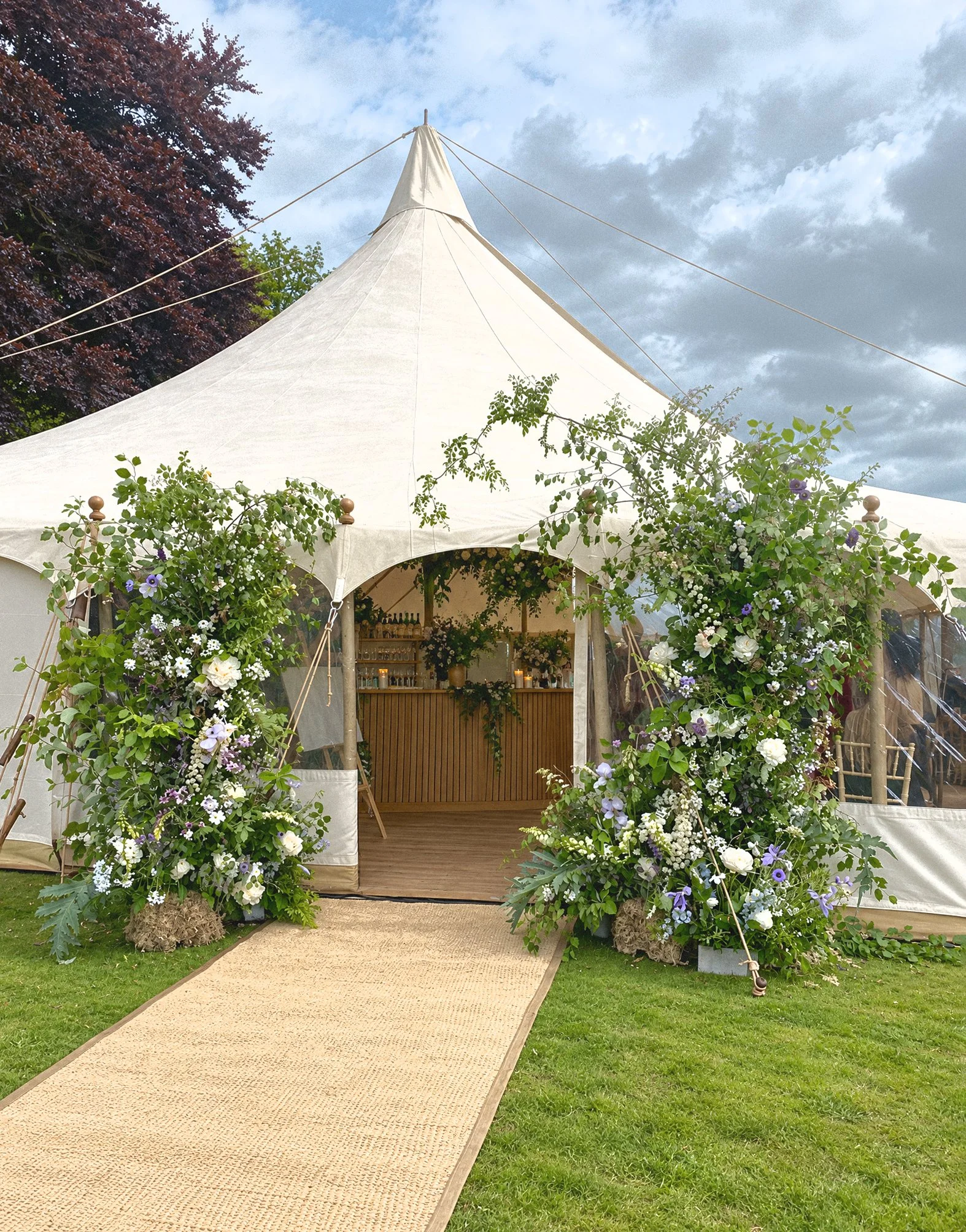 A large white event tent decorated with lush green and purple floral arrangements at the entrance. A beige carpet walkway leads up to the tent on a grassy area with trees and a cloudy sky overhead.