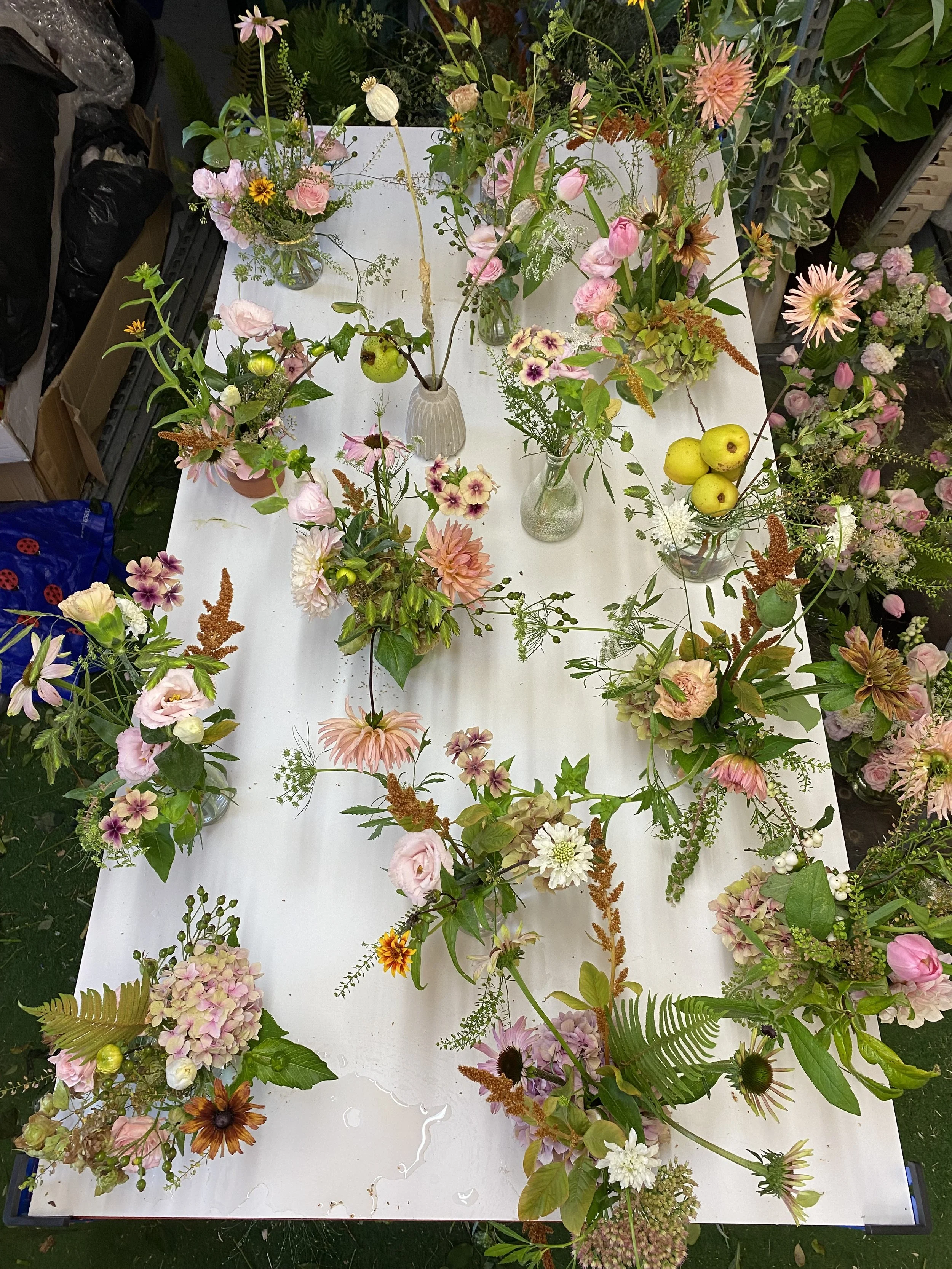 Various fresh flowers and some apples arranged in vases and laid on a white table.