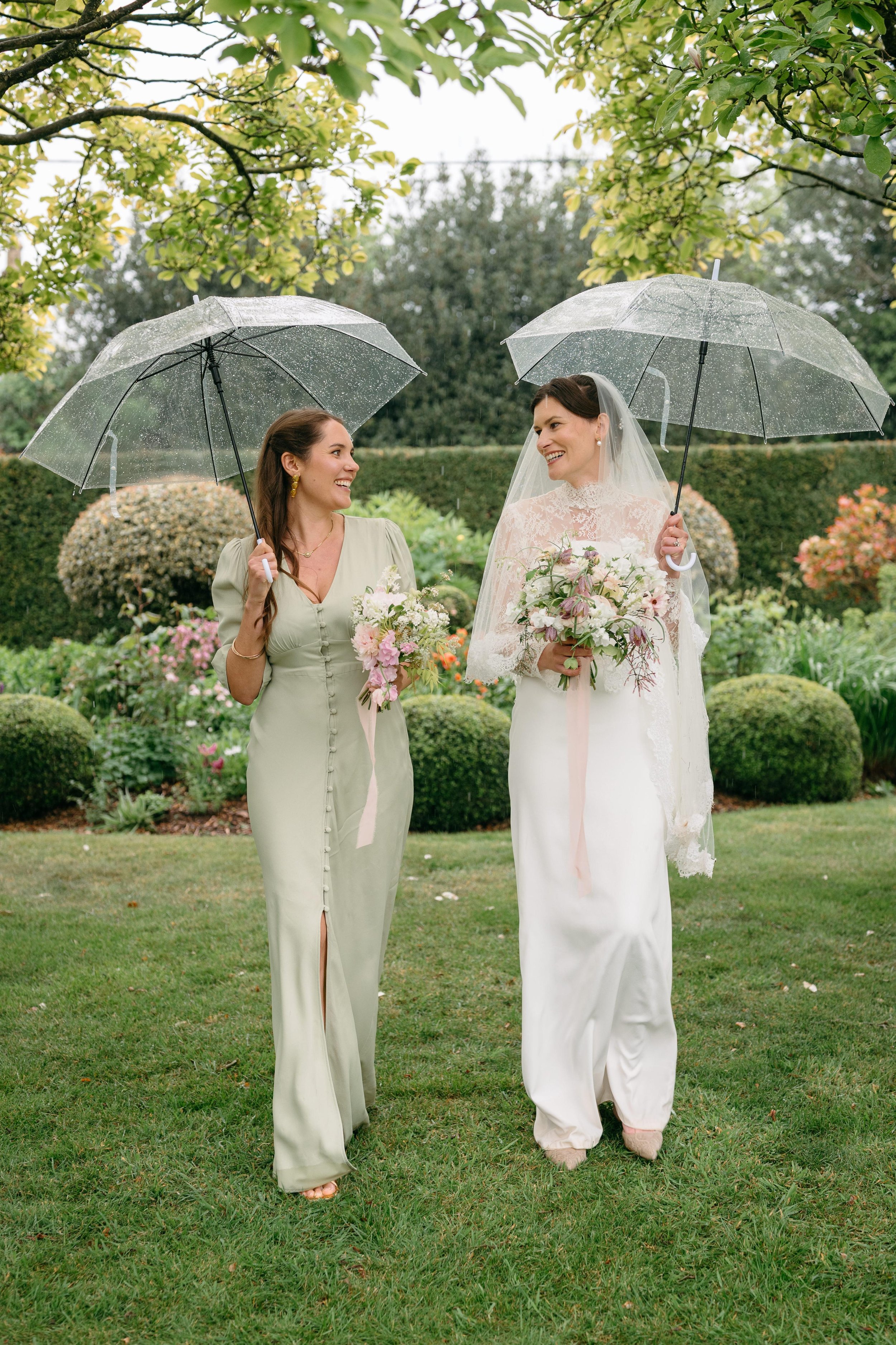 Two women standing outdoors in a garden, smiling and holding clear umbrellas, with one woman dressed as a bride in a white wedding gown and veil, and the other in a light green dress, both carrying bouquets of flowers.