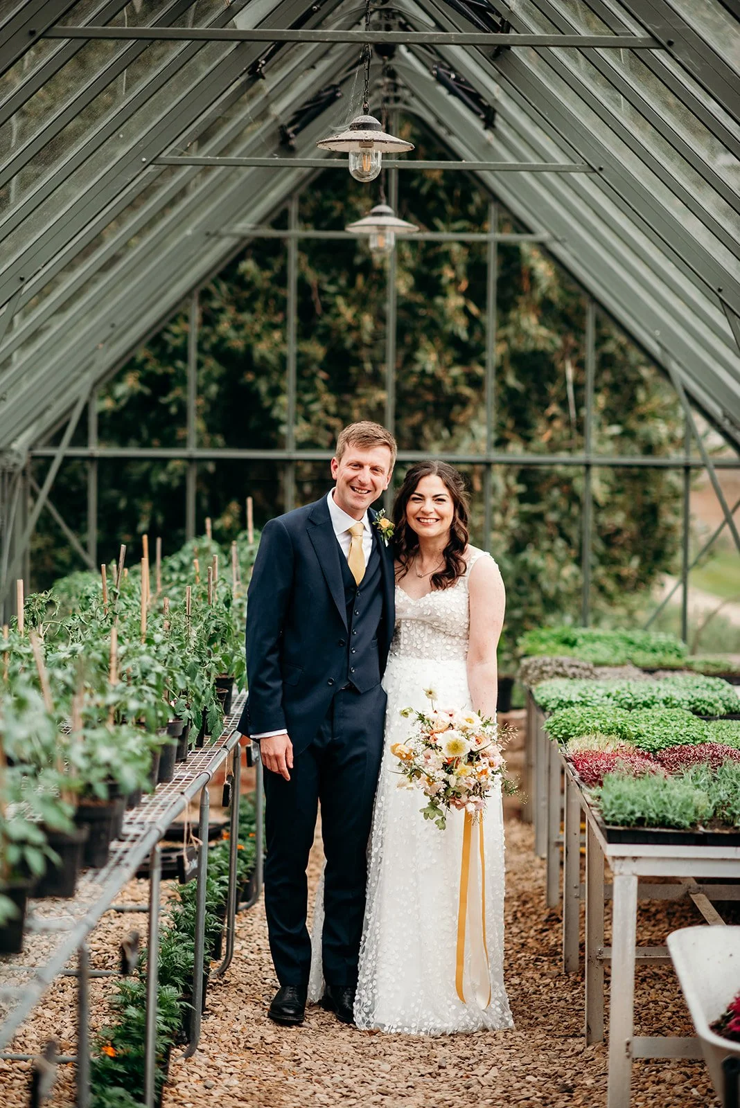 A happy newlywed couple posing inside a greenhouse, with green plants in pots on both sides, the bride holding a bouquet of flowers, and the groom wearing a dark suit and yellow tie.