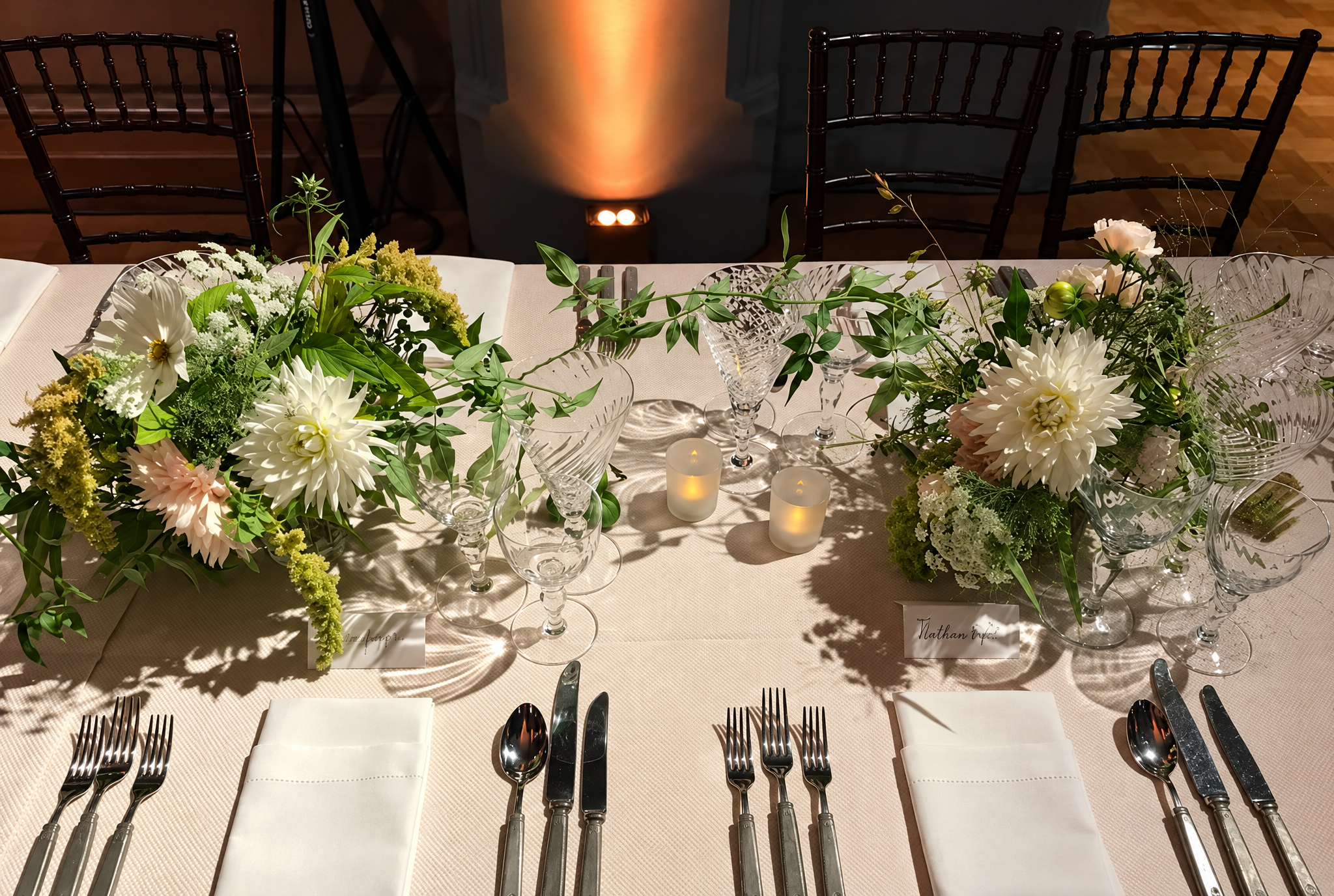 A formal dining table decorated with white and green floral centerpieces, surrounded by silverware, white napkins, glassware, and small candles, set for an event or celebration.