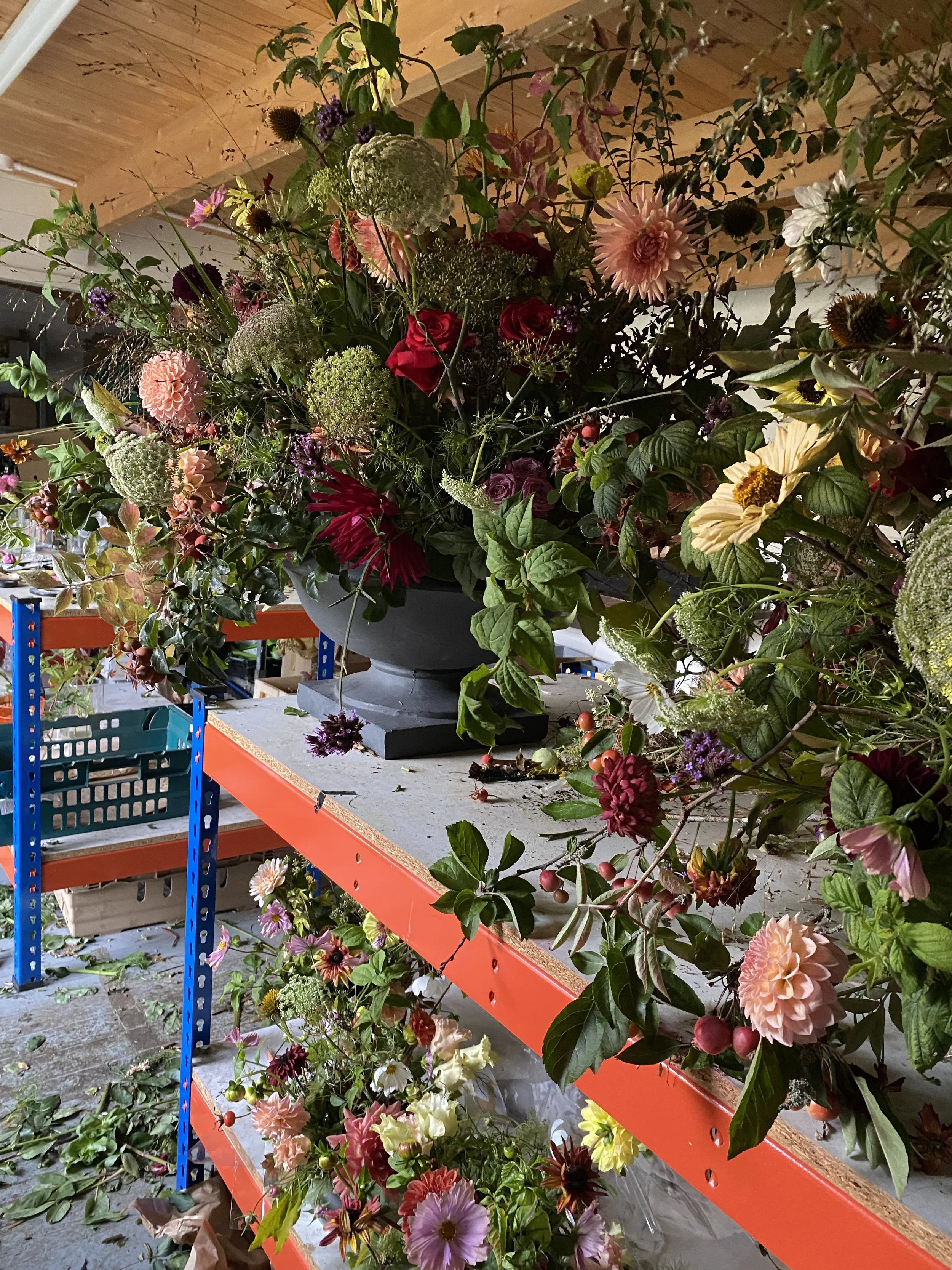 Large colorful flower arrangement on a metal table in a workshop or storage room with shelves and stained wooden ceiling.