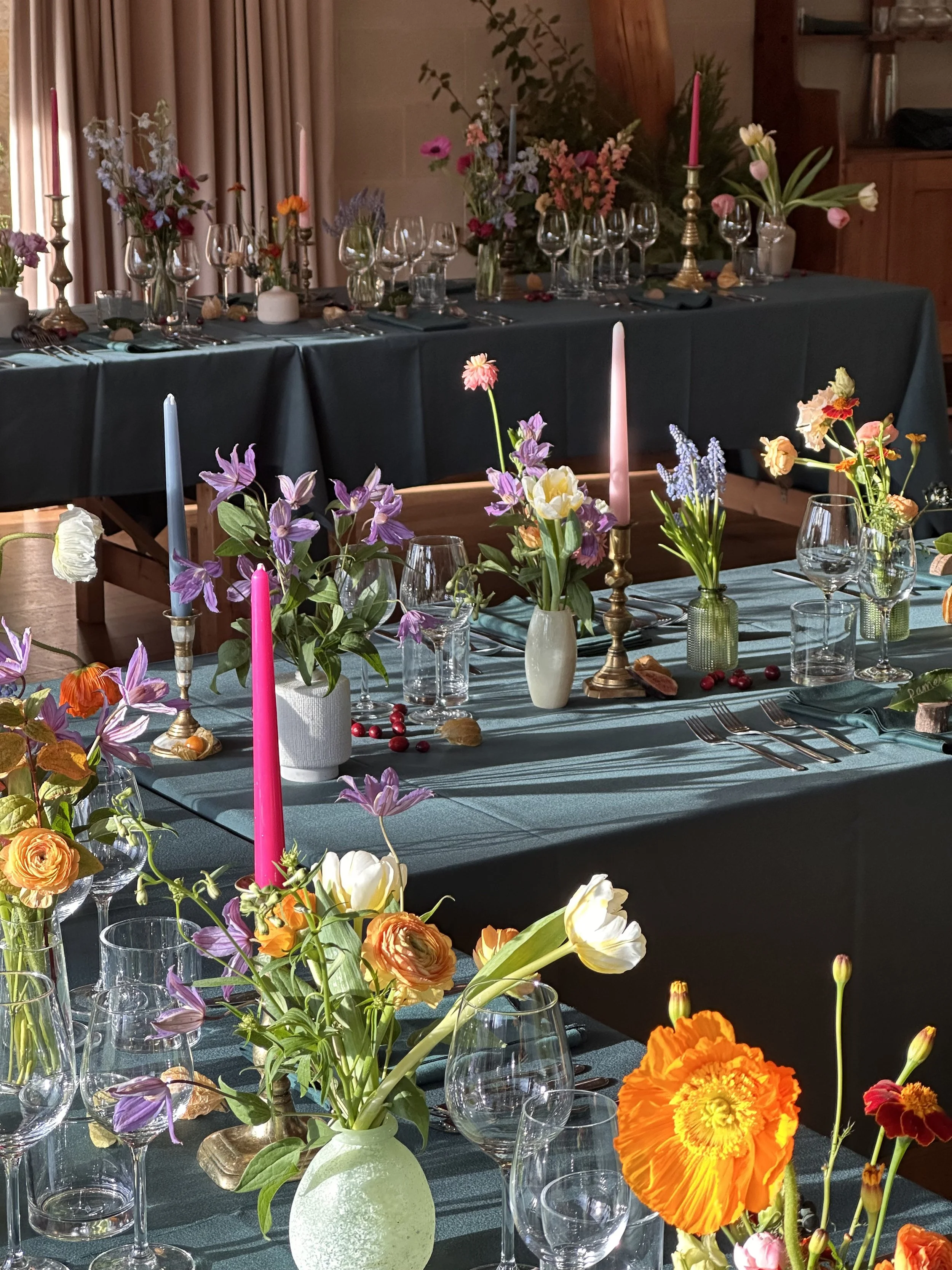 Elegant dining table set with flower arrangements, candles, wine glasses, and cutlery in a room with curtains and wooden accents.