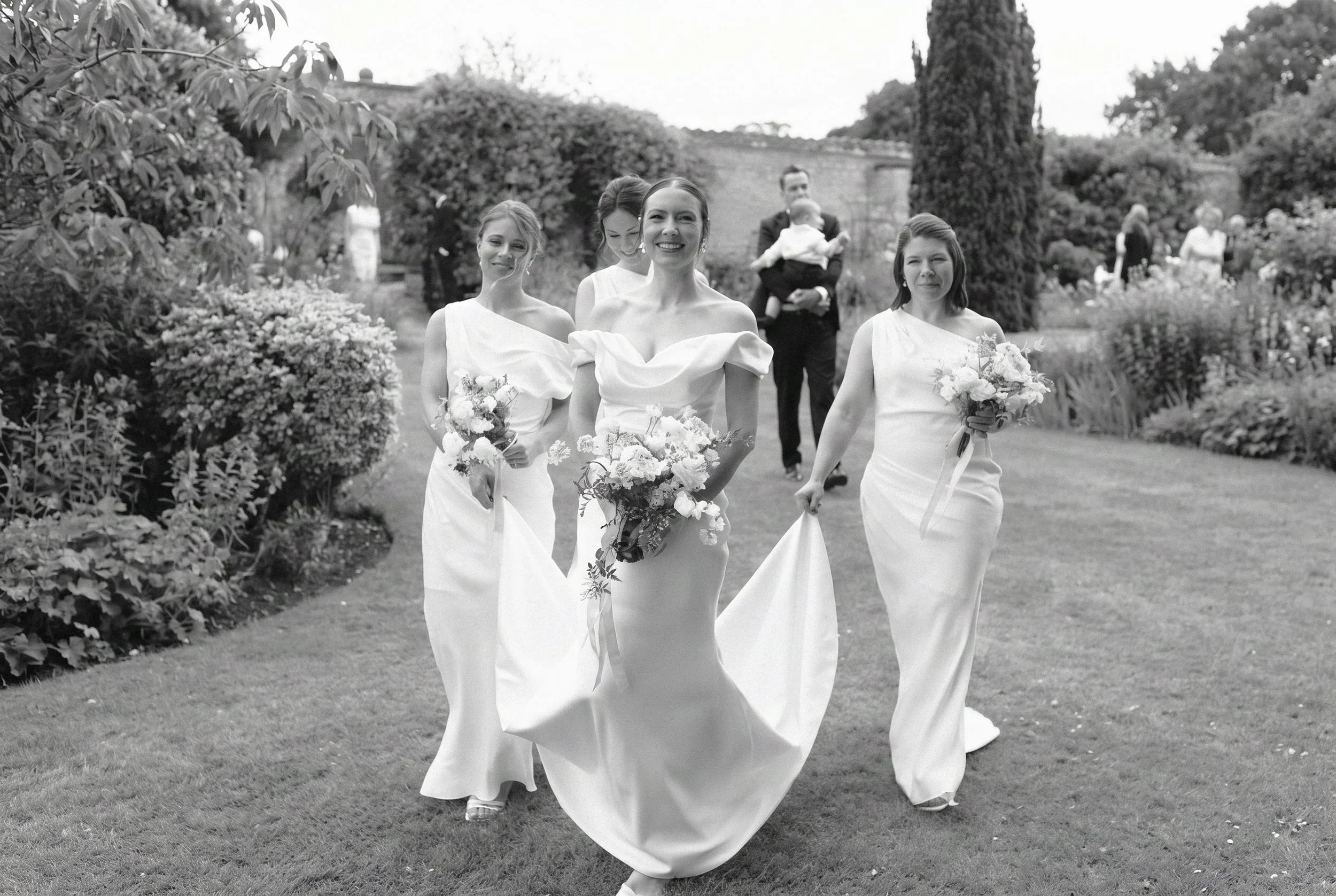 Four women in white gowns walking through a garden during a wedding, with a man in a suit holding a child in the background.