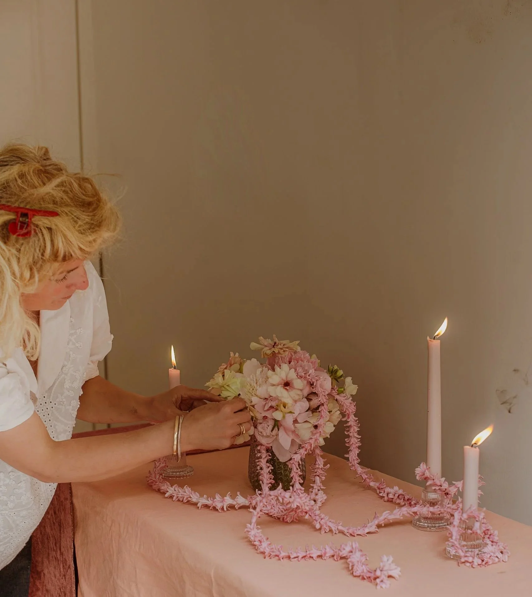 Woman arranging pink and white flower bouquet on table with pink floral lei, candles, and flower petals.
