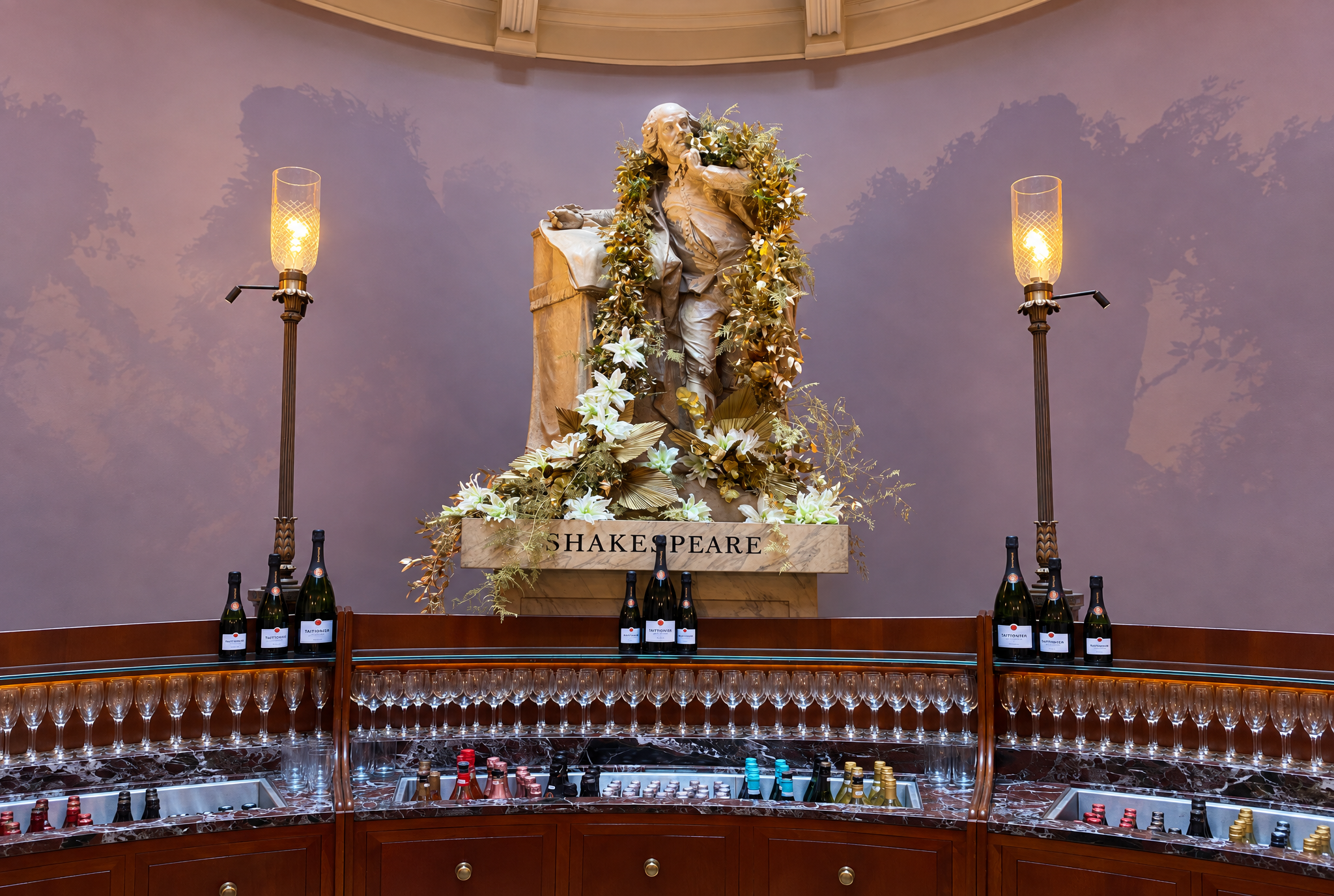 Statue of William Shakespeare adorned with a floral garland, situated in a room with a purple wall, flanked by two wall lamps. In front of the statue, there are bottles of sparkling wine and glasses on a marble shelf, with an array of champagne bottles and glasses below.