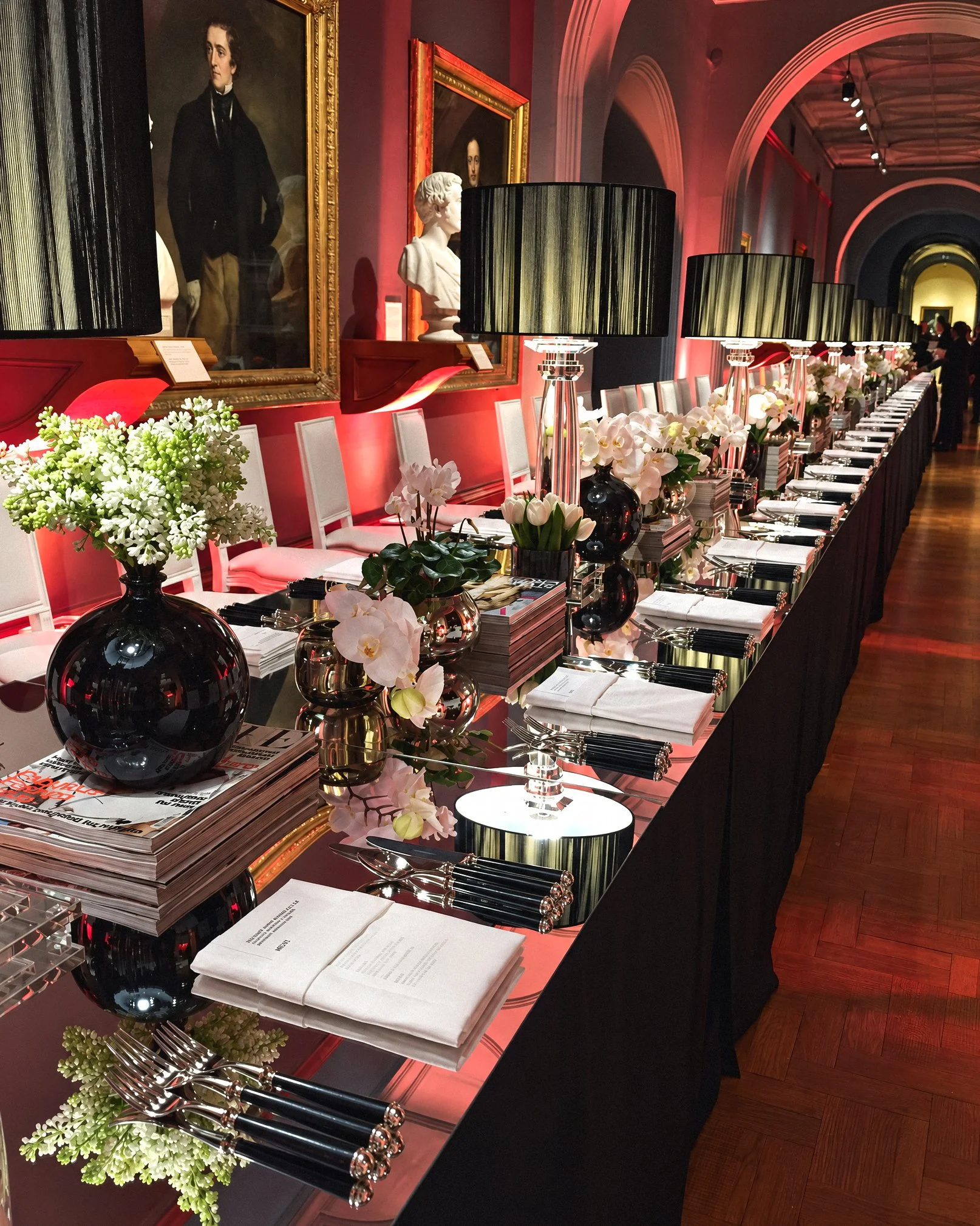 A long banquet table set for a formal event in a grand hall with high ceilings, pink walls, and large framed portraits of historical figures. The table is decorated with black lamps, white floral arrangements, and neatly arranged silverware, napkins, and newspapers.