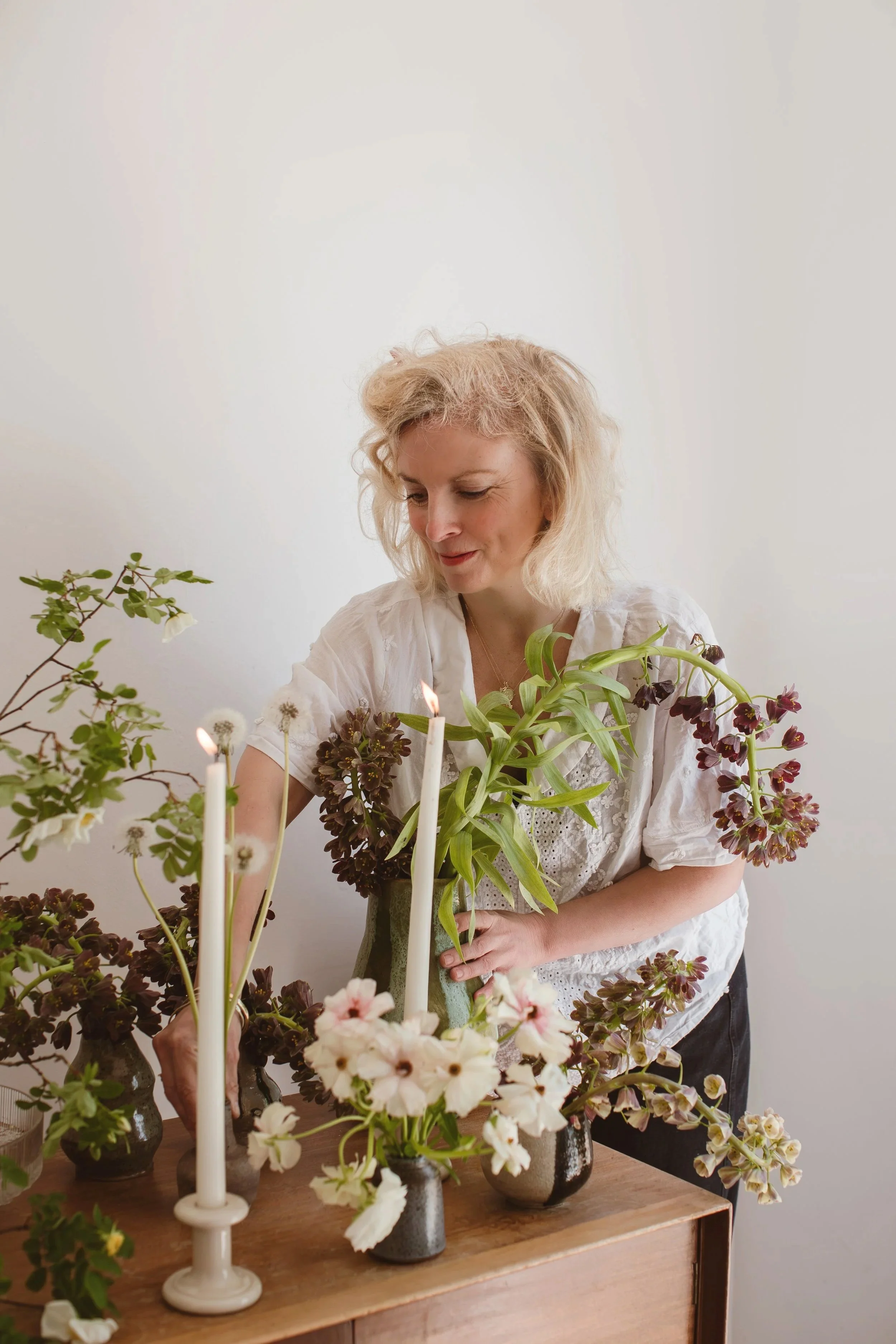A woman with curly blonde hair arranging flowers on a wooden table, surrounded by several vases with different flowers and a white candlestick, against a plain off-white wall.
