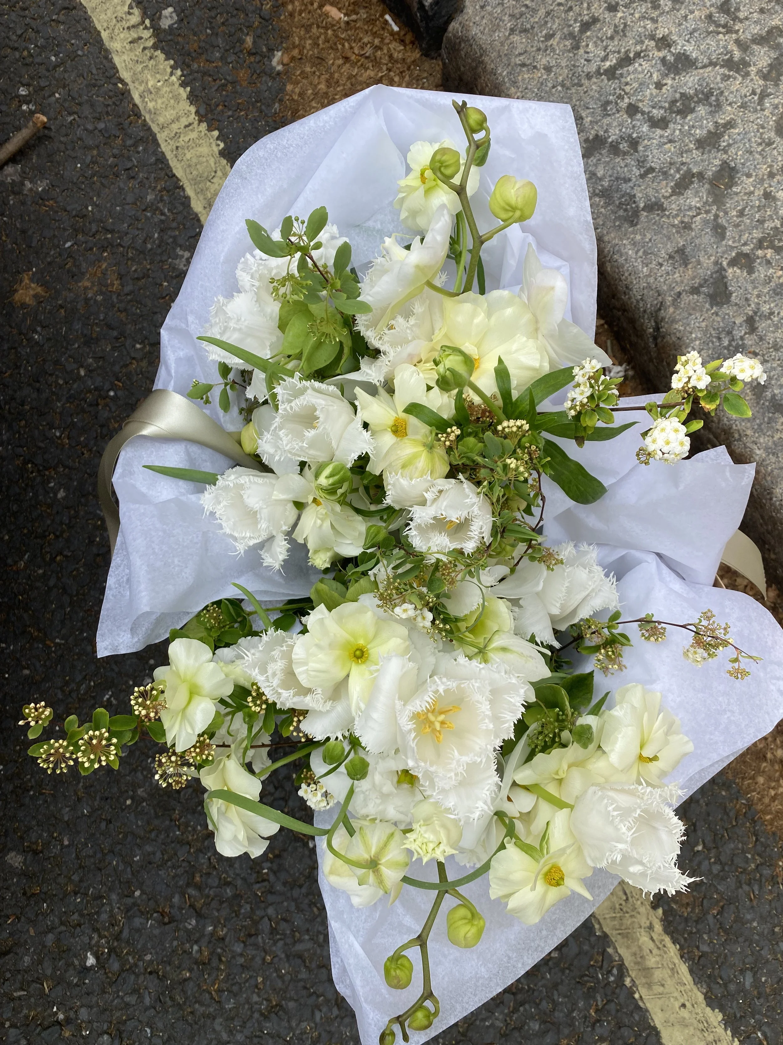 A bouquet of white flowers with green leaves wrapped in white paper is placed on the ground next to a curb and asphalt road.