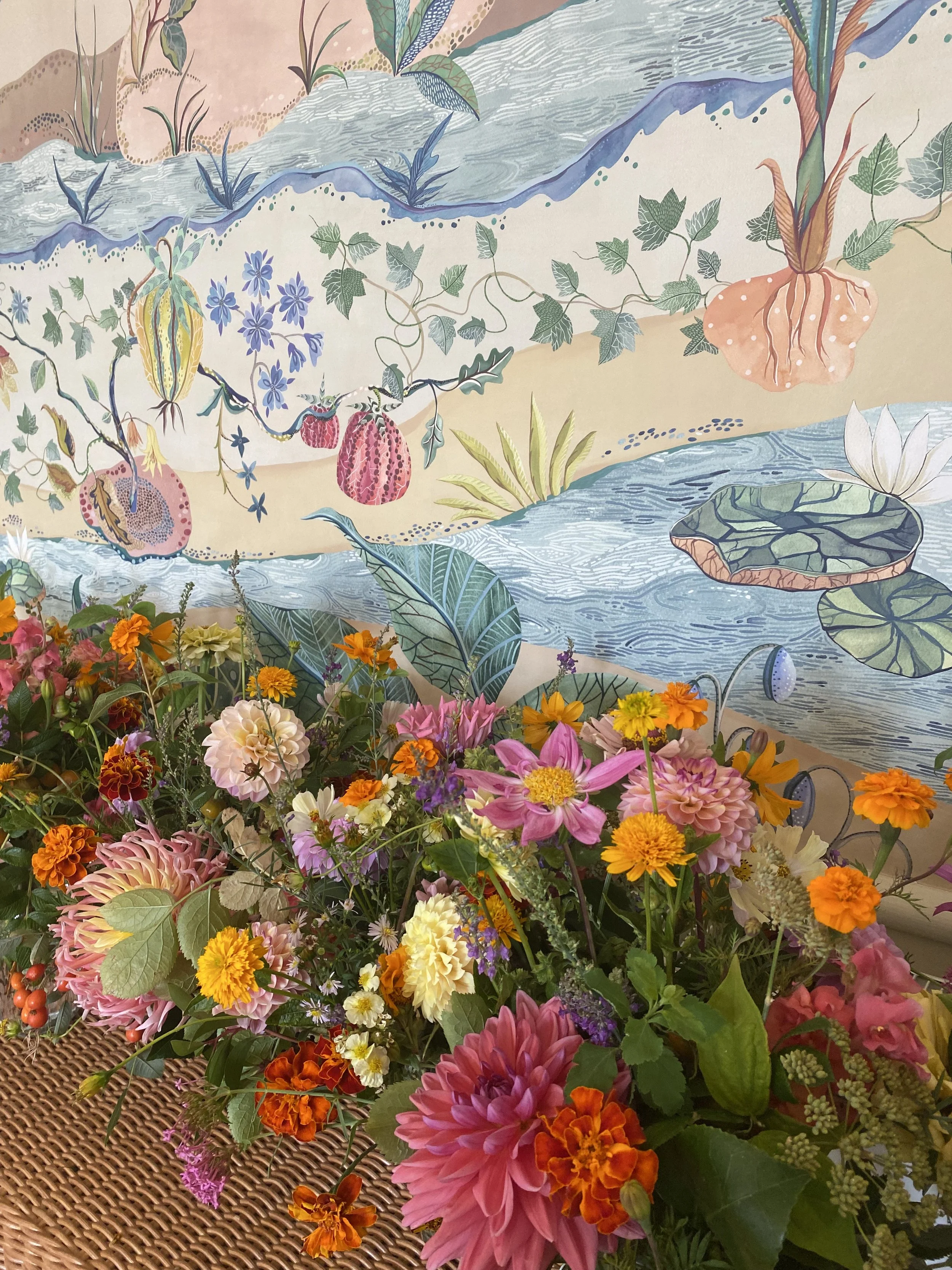 Colorful flower arrangement with various types of flowers and greenery in front of a decorative wall with a mural of water, plants, and abstract patterns.