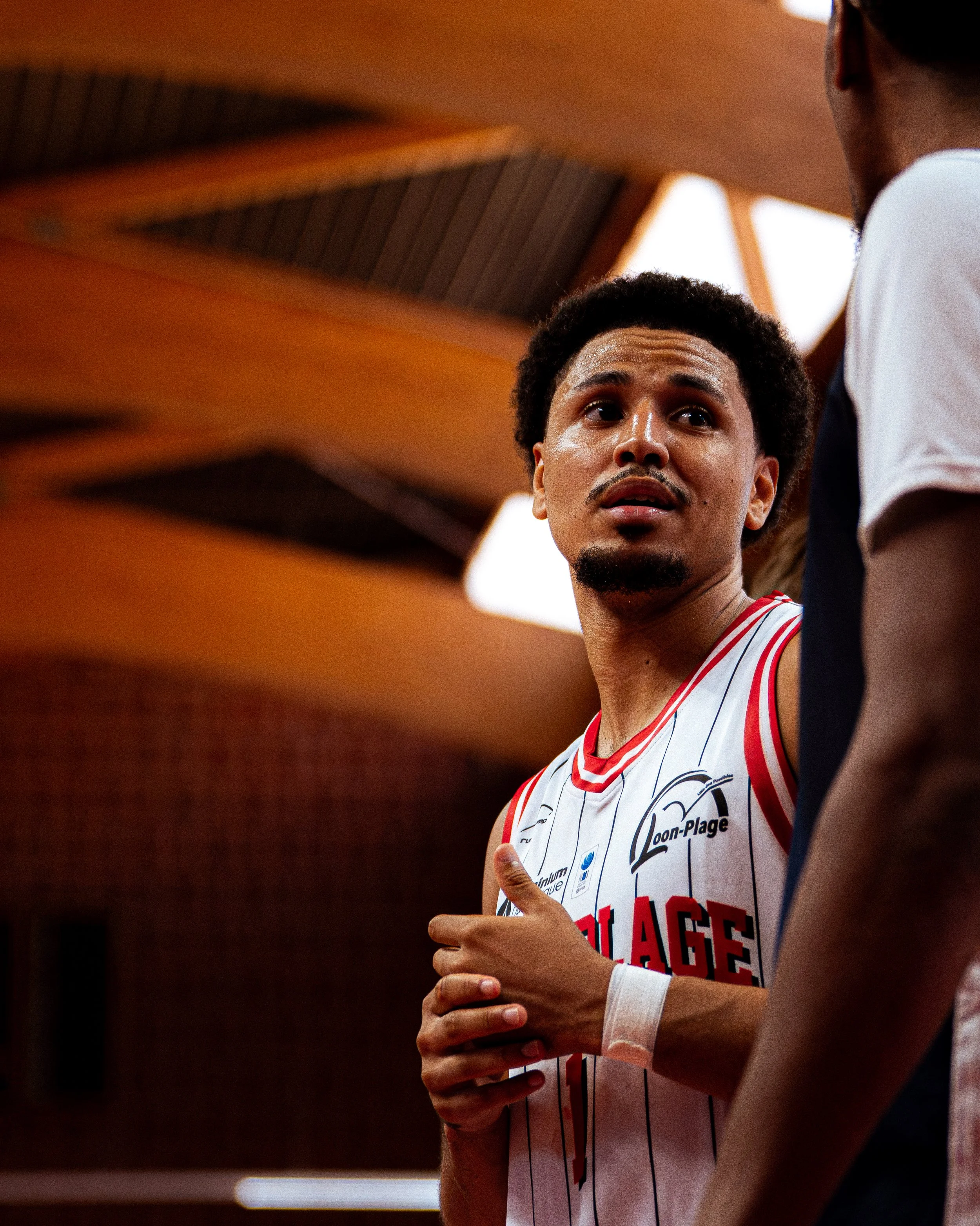 Deux hommes, dont un porte un maillot de basketball avec le nom Chicago, discutent dans une salle intérieure. Le maillot a des rayures verticales et un logo "Loon-Plage".