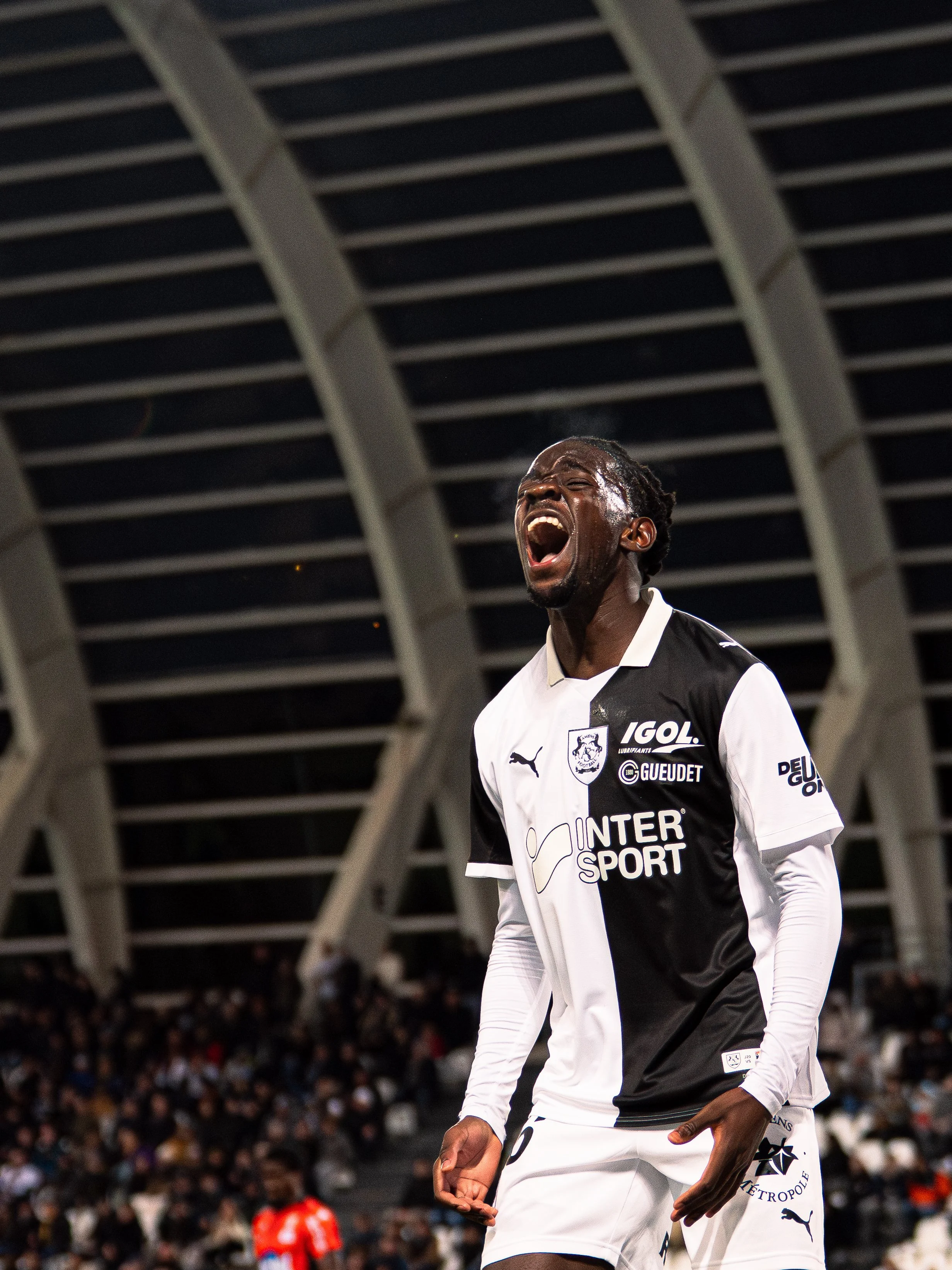 Un joueur de football en train de crier, portant un maillot blanc et noir avec logos et sponsors, dans un stade rempli de spectateurs.
