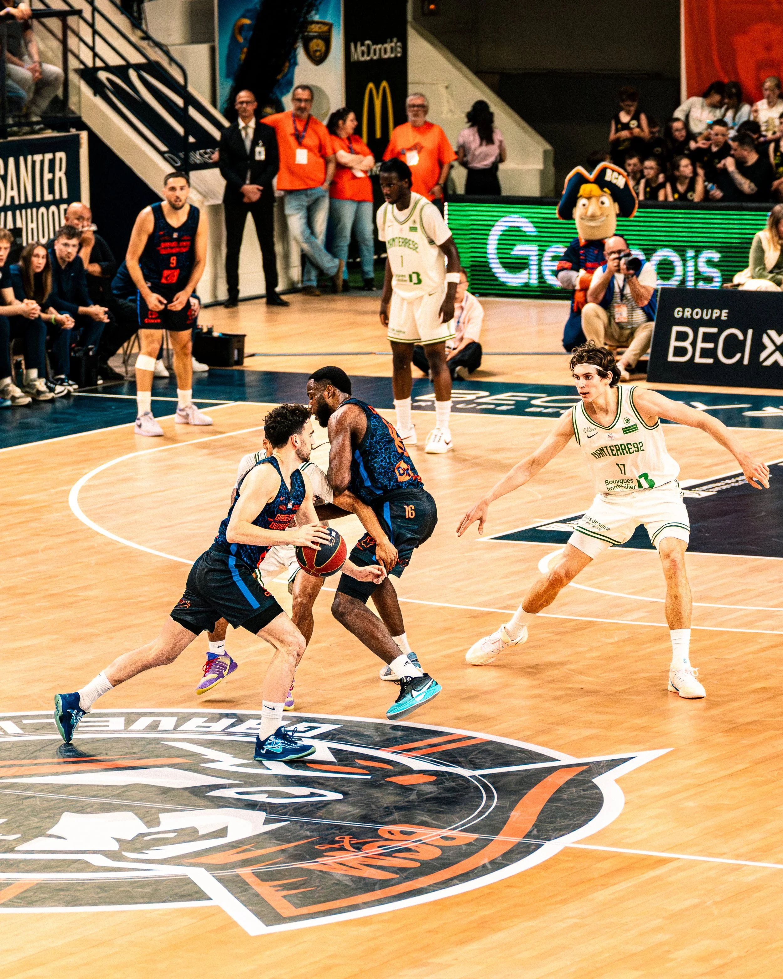 Match de basketball avec des joueurs en action, un arbitre et une mascotte sur le côté droit.