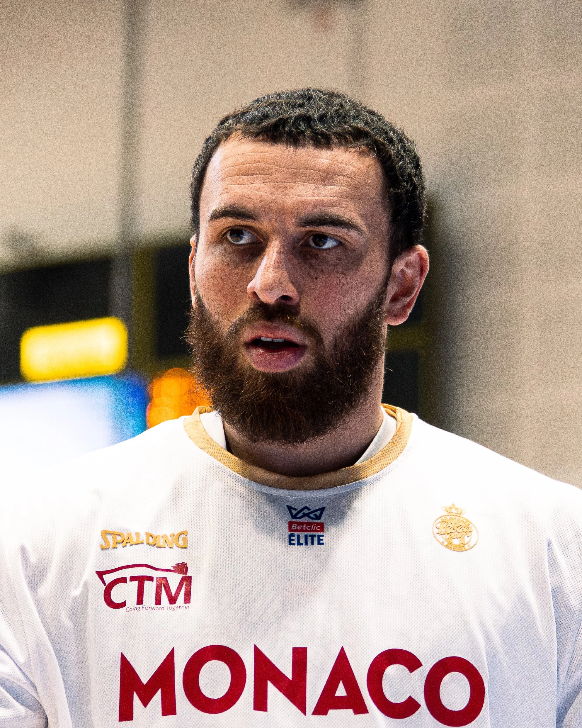 Portrait d'un homme avec une barbe, portant un maillot de hockey blanc orné de plusieurs logos et modules. Il semble être au centre d'une conversation ou d'une interview.