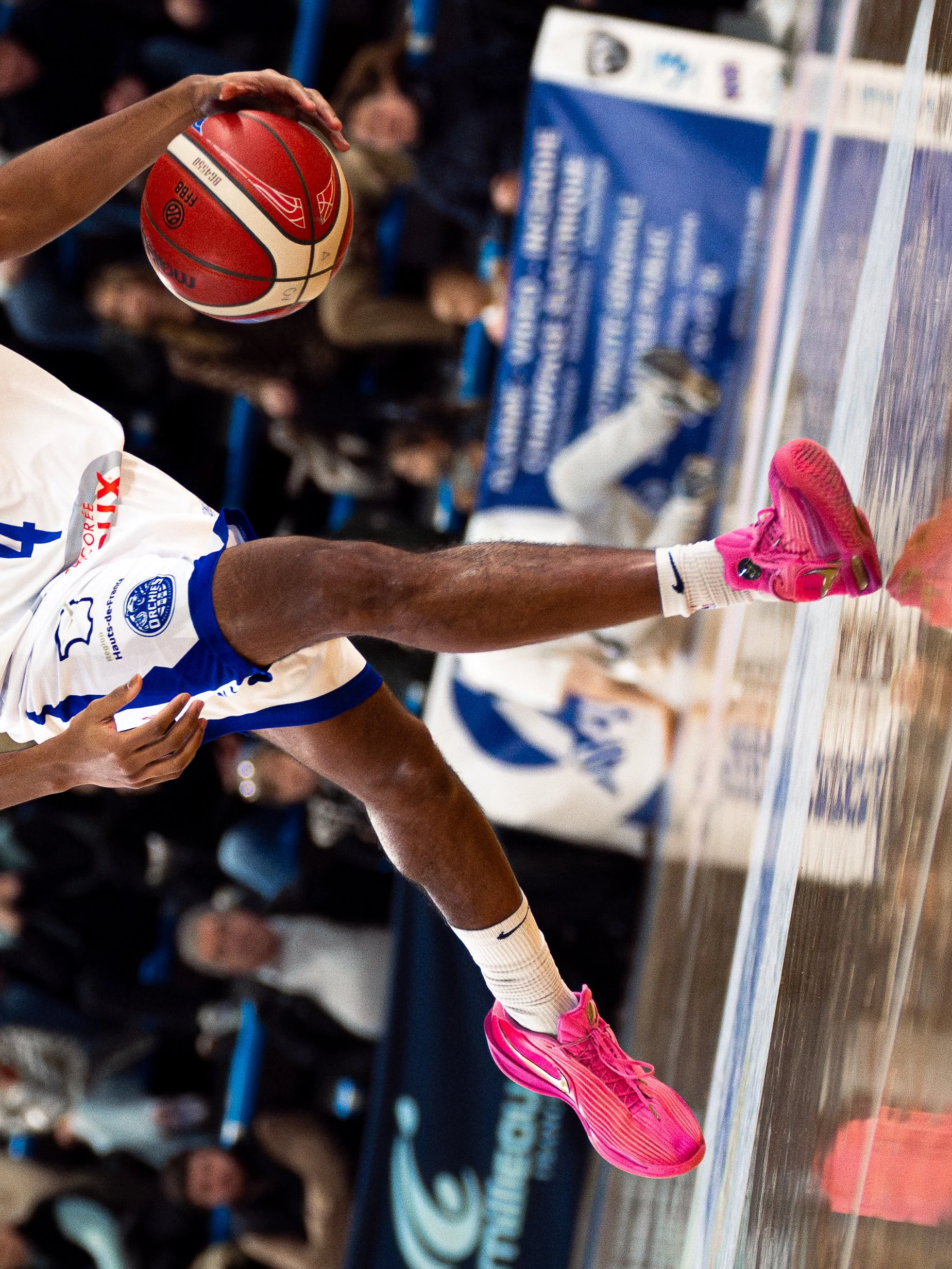 Un joueur de basketball en tenue blanche et roses effectue un dunk lors d'un match. Le sol du court est en bois, avec un public en arrière-plan.