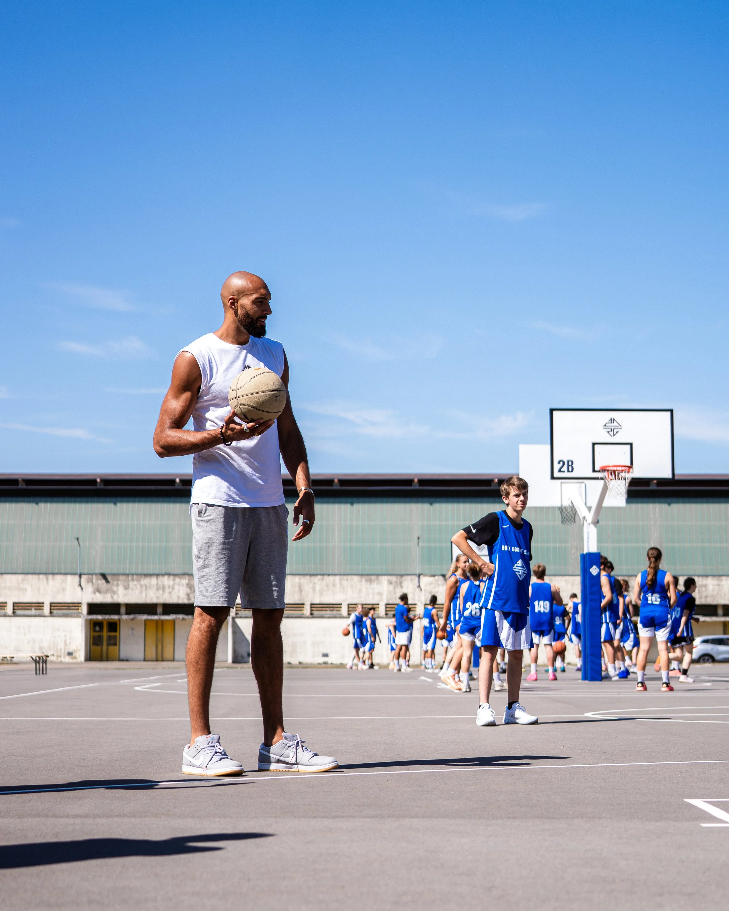 Un entraîneur masculin tien un ballon de basket sur un terrain extérieur de basketball, avec un groupe d'enfants en uniforme de basketball bleu en arrière-plan. Le ciel est clair et ensoleillé.