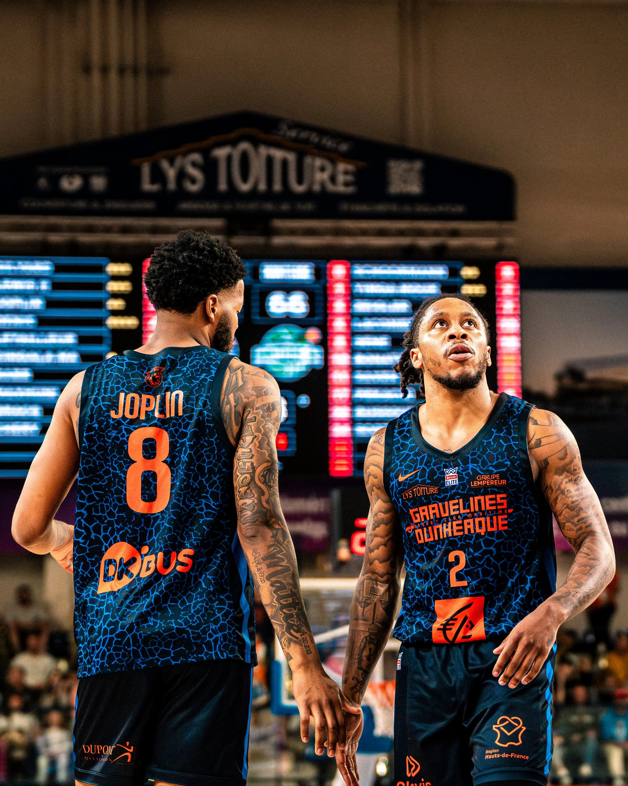 Deux joueurs de basket-ball en uniforme regardant vers le haut lors d'un match, un dans le dos avec le nom 'JOPLIN' et le numéro 8, l'autre à l'avant avec le numéro 2, dans une salle de sport.
