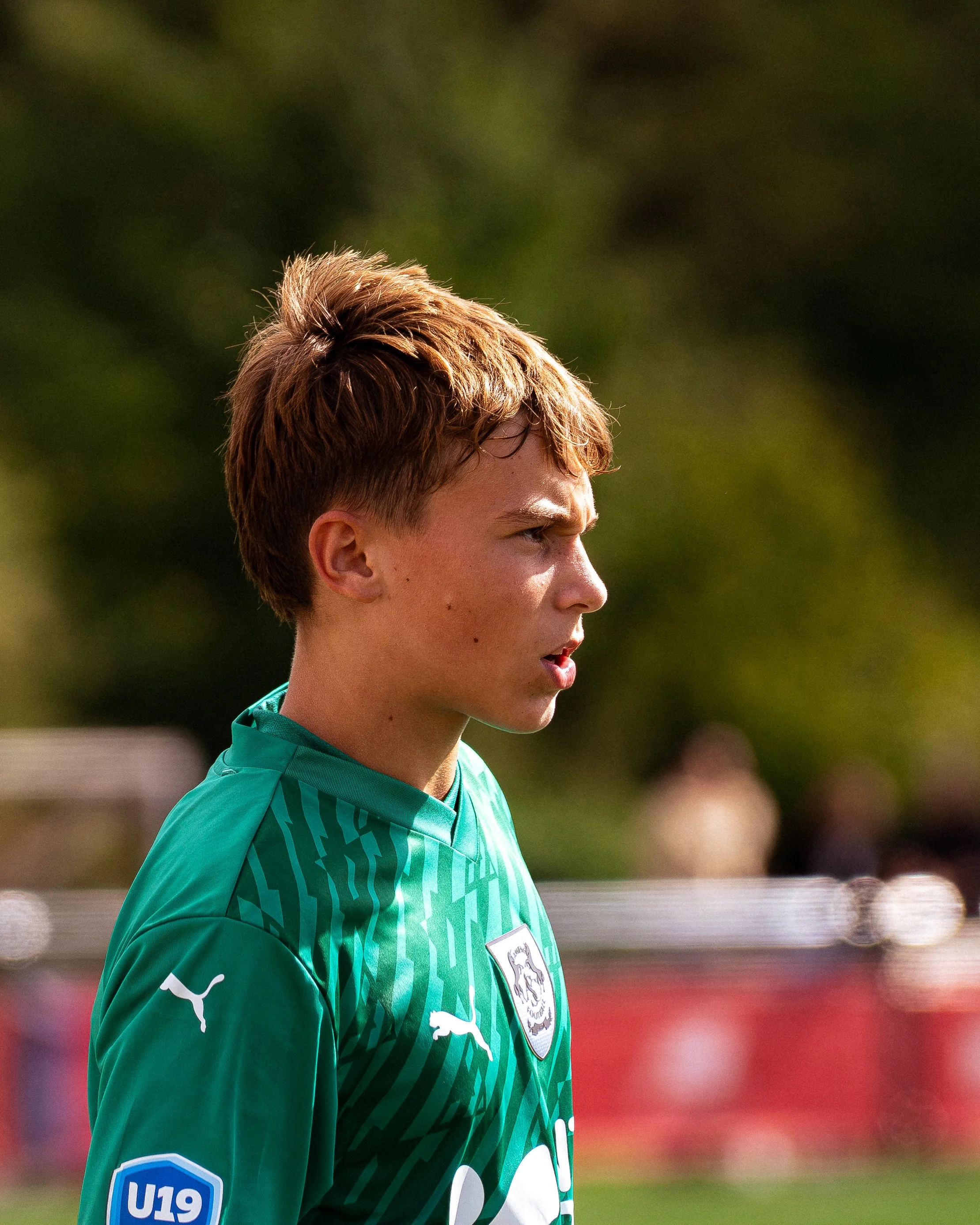 Jeune homme avec cheveux courts et roux portant un maillot vert de football avec un logo de la Fédération allemande et un badge U19, regardant vers la droite, en extérieur lors d'un match.