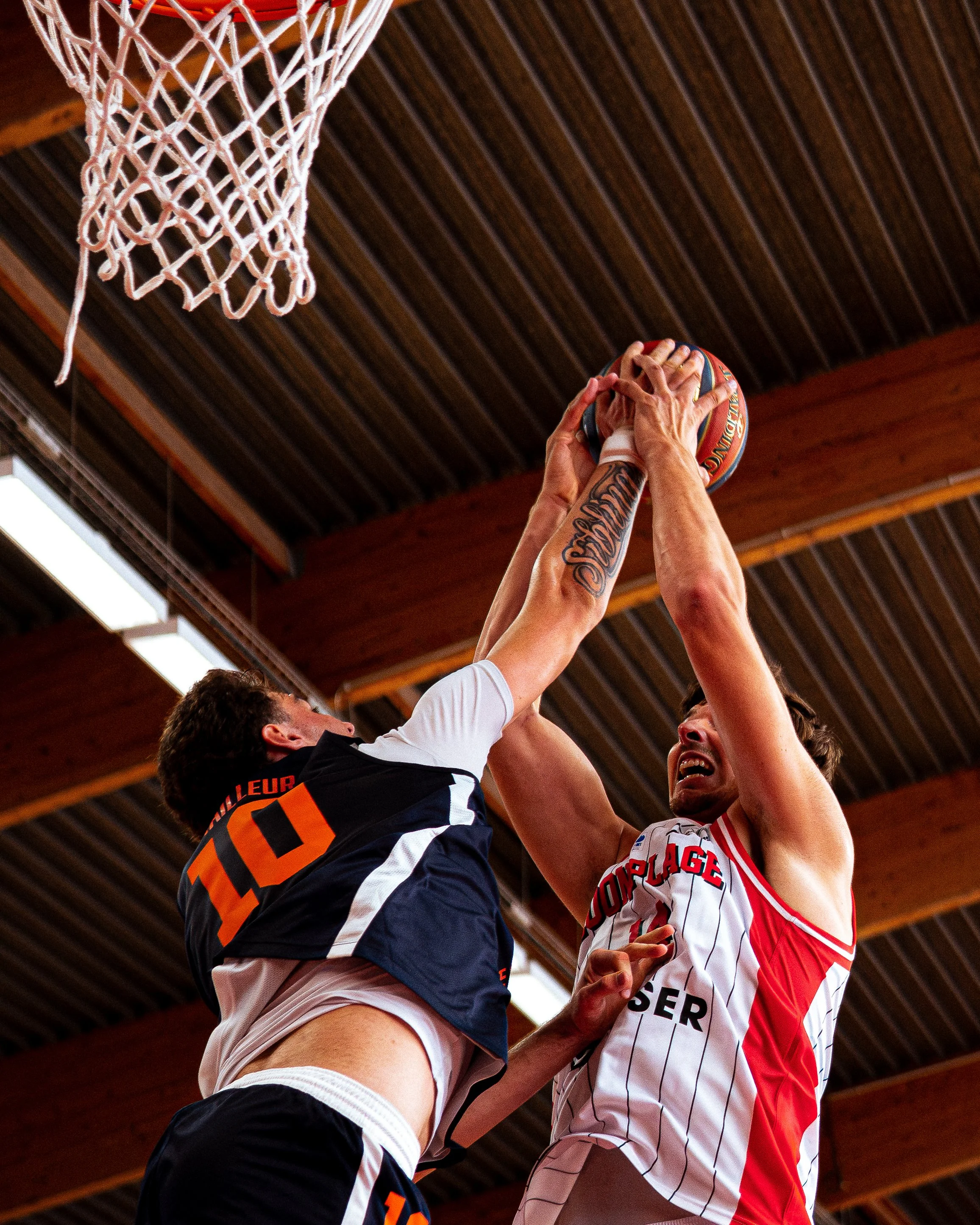 Deux basketballs sont en train d'être contestés par deux joueurs lors d'un match, sous un plafond en bois.