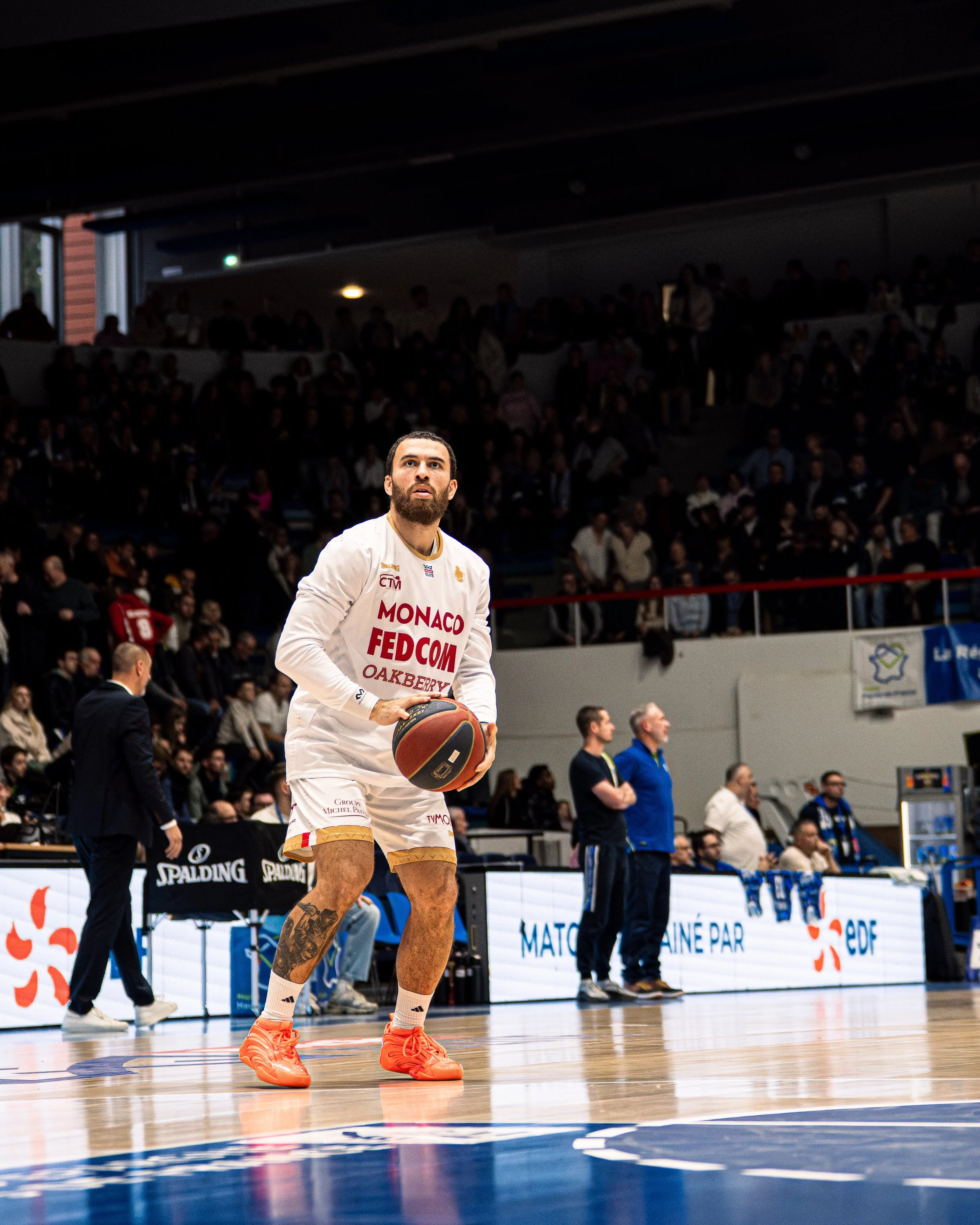 Un joueur de basketball en tenue blanche avec le mot Monaco sur le maillot, tenant un ballon de basket, sur un terrain de basketball lors d'un match, avec un public en arrière-plan.